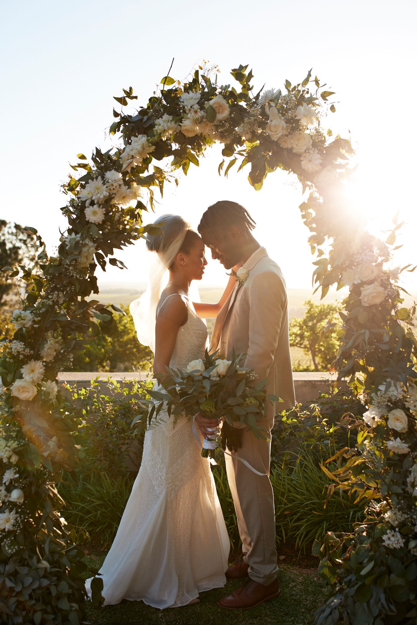 Couple stands under floral arch, bathed in sunlight, at outdoor wedding.