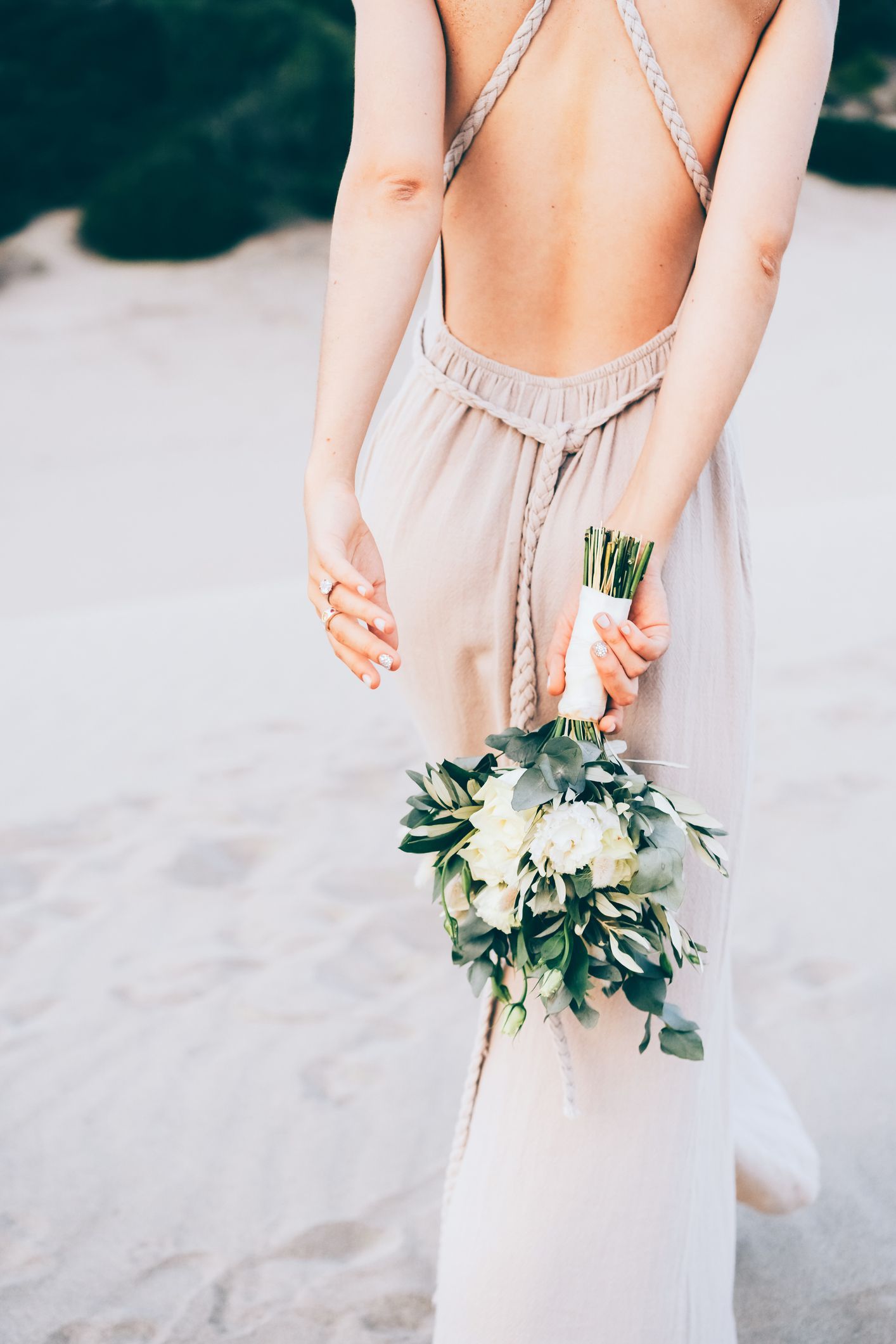 Woman in backless dress, holding a white and green bouquet, standing on sand.