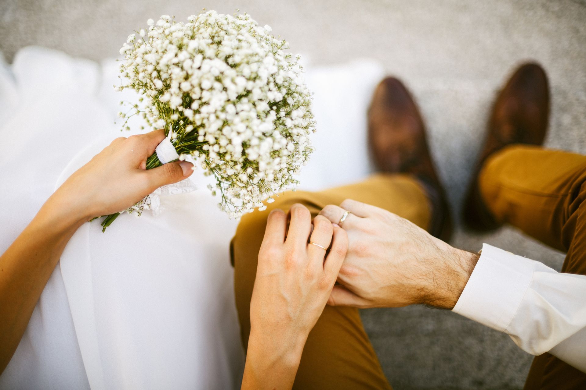 Bride holding bouquet, rests hand on groom’s as they sit. Groom wears brown shoes and pants.
