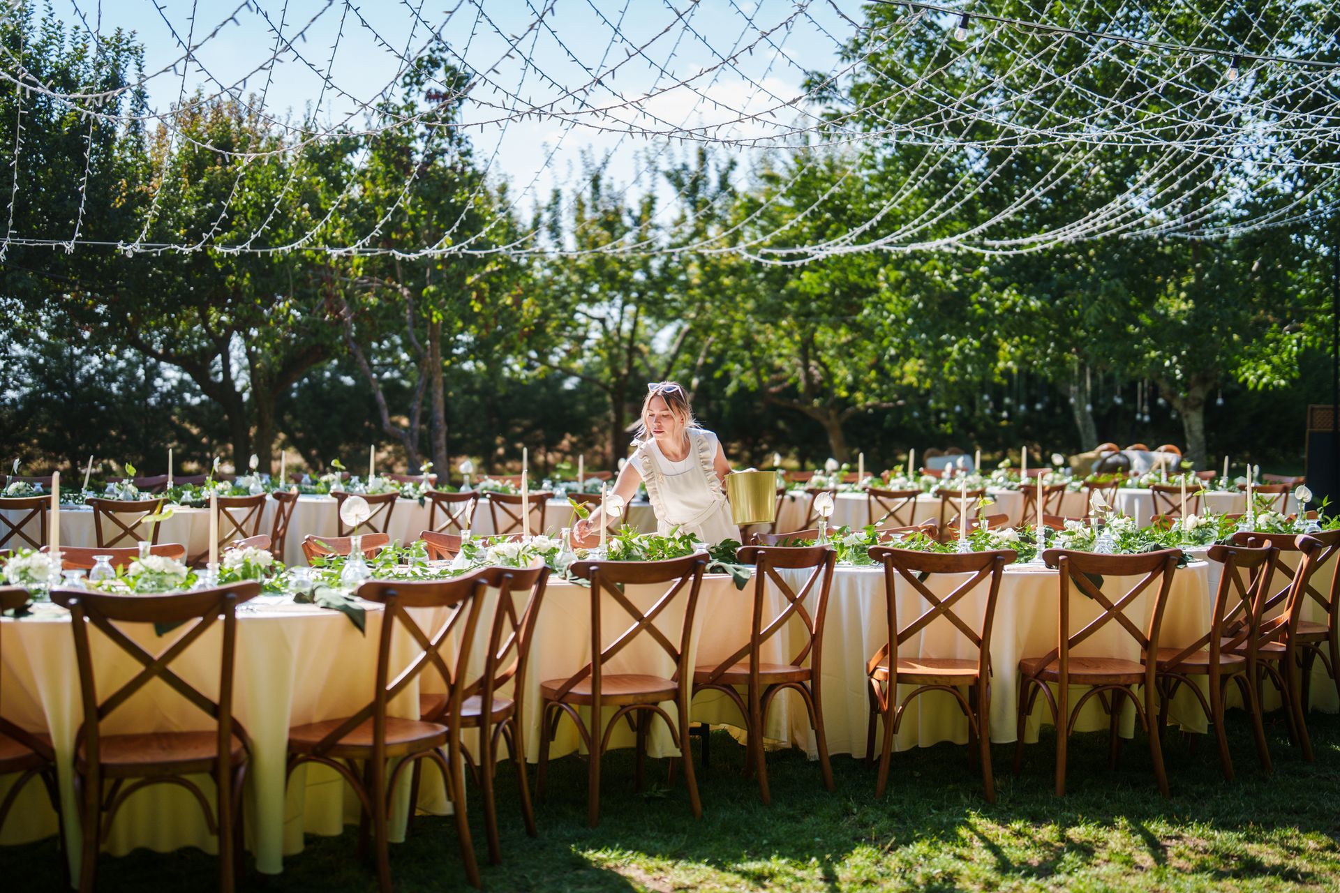 Outdoor wedding reception setup with tables, chairs, and decorations. Person arranges flowers on a table.