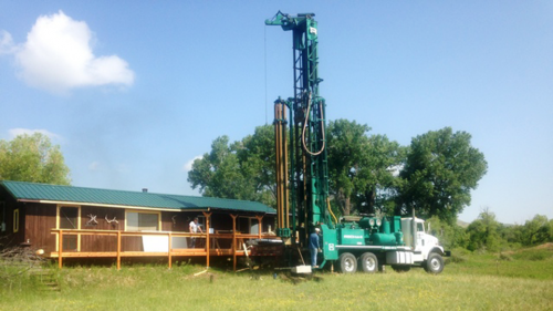 A large teal drilling rig is parked on a grassy field next to a brown cabin with a green roof under a clear blue sky.