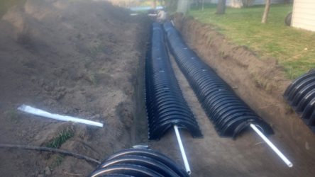 Two rows of black, arched plastic drainage chambers installed in a deep trench, connected by white PVC pipes.