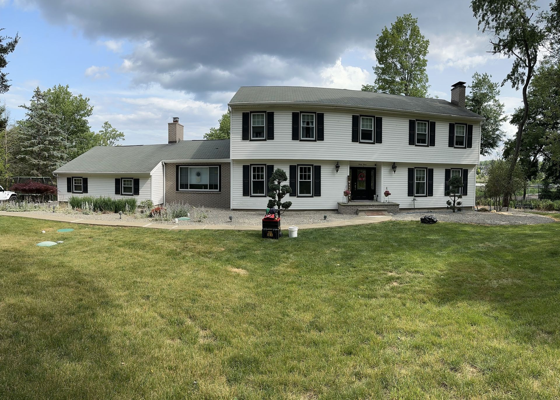 White two-story farmhouse with black shutters and stone foundation on a grassy lawn under a cloudy sky