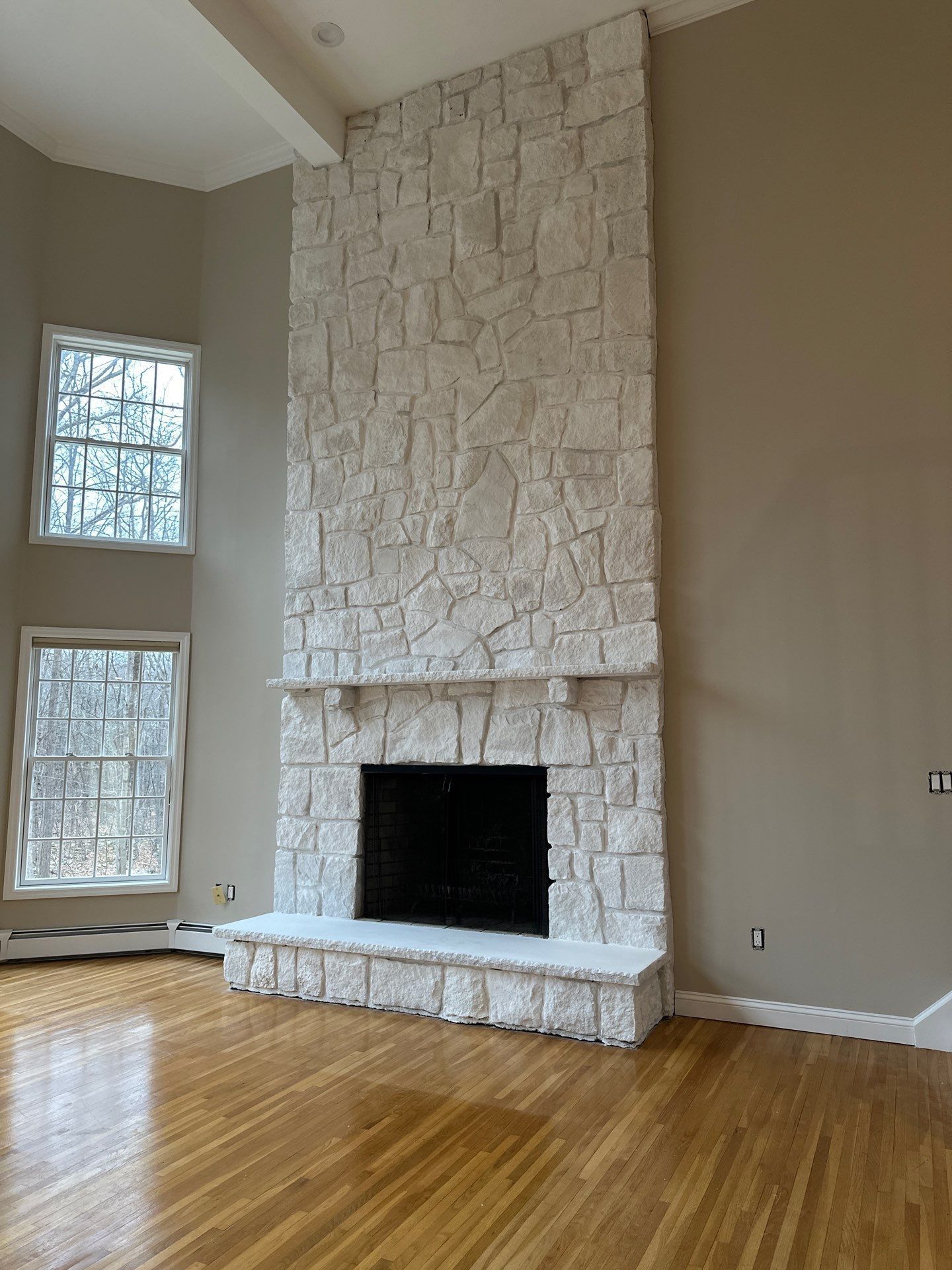 Stone fireplace, reaching high ceiling. Two windows to the left. Wooden floor and tan walls.