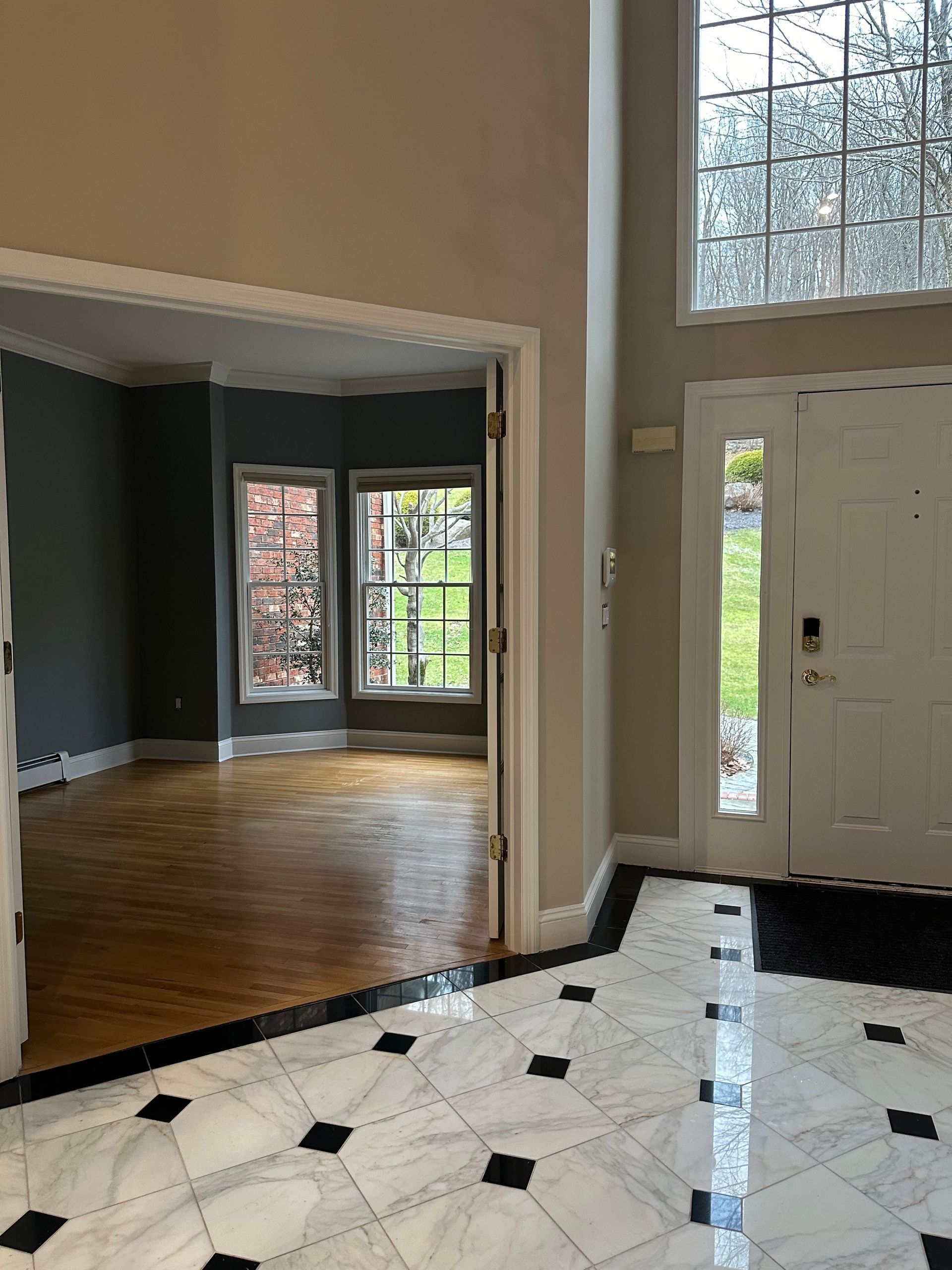 Entryway with marble tile floor, door to the outside, and view into a room with windows and hardwood floors.