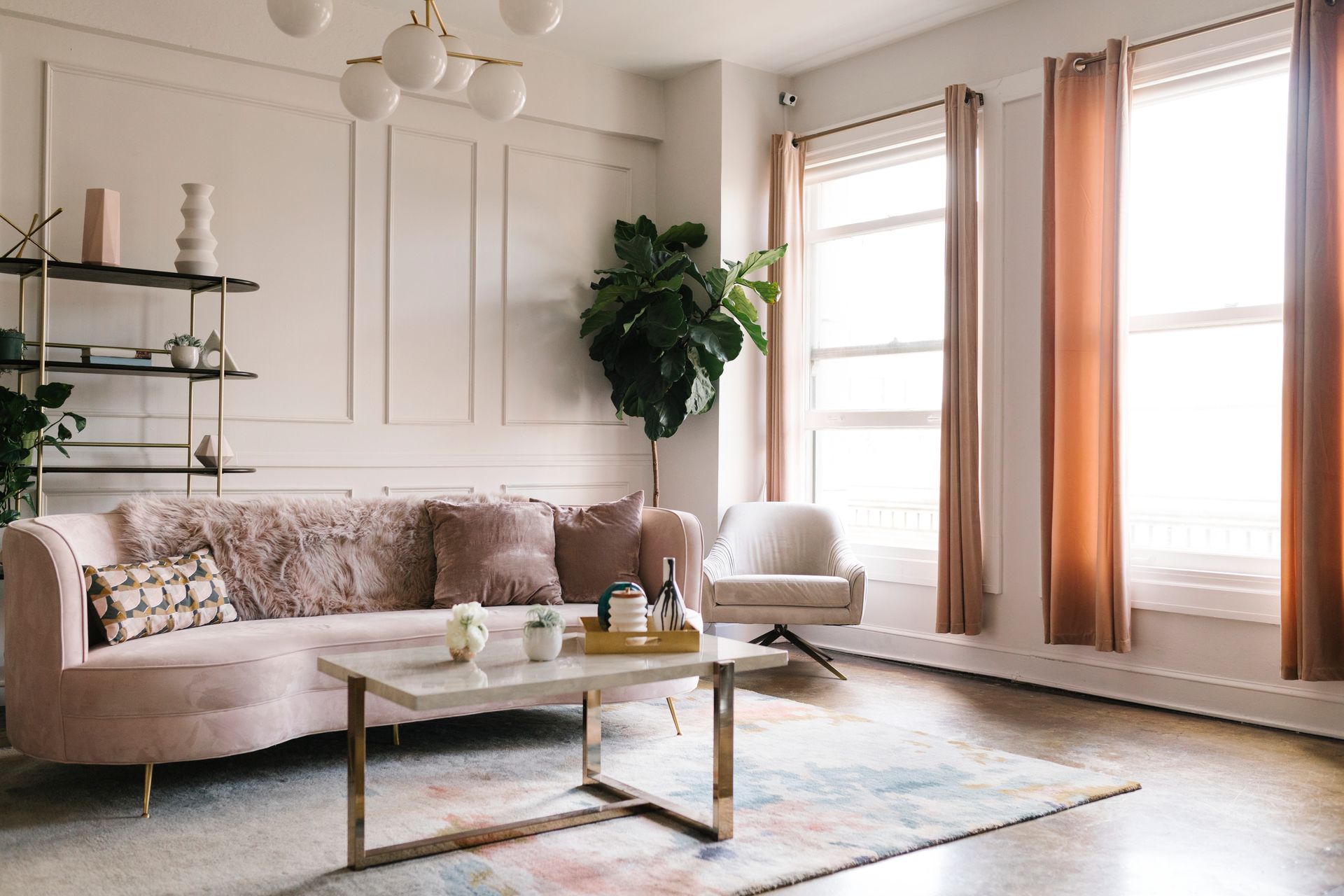 Bright living room with beige sofa, coffee table, plants, and large windows with warm curtains