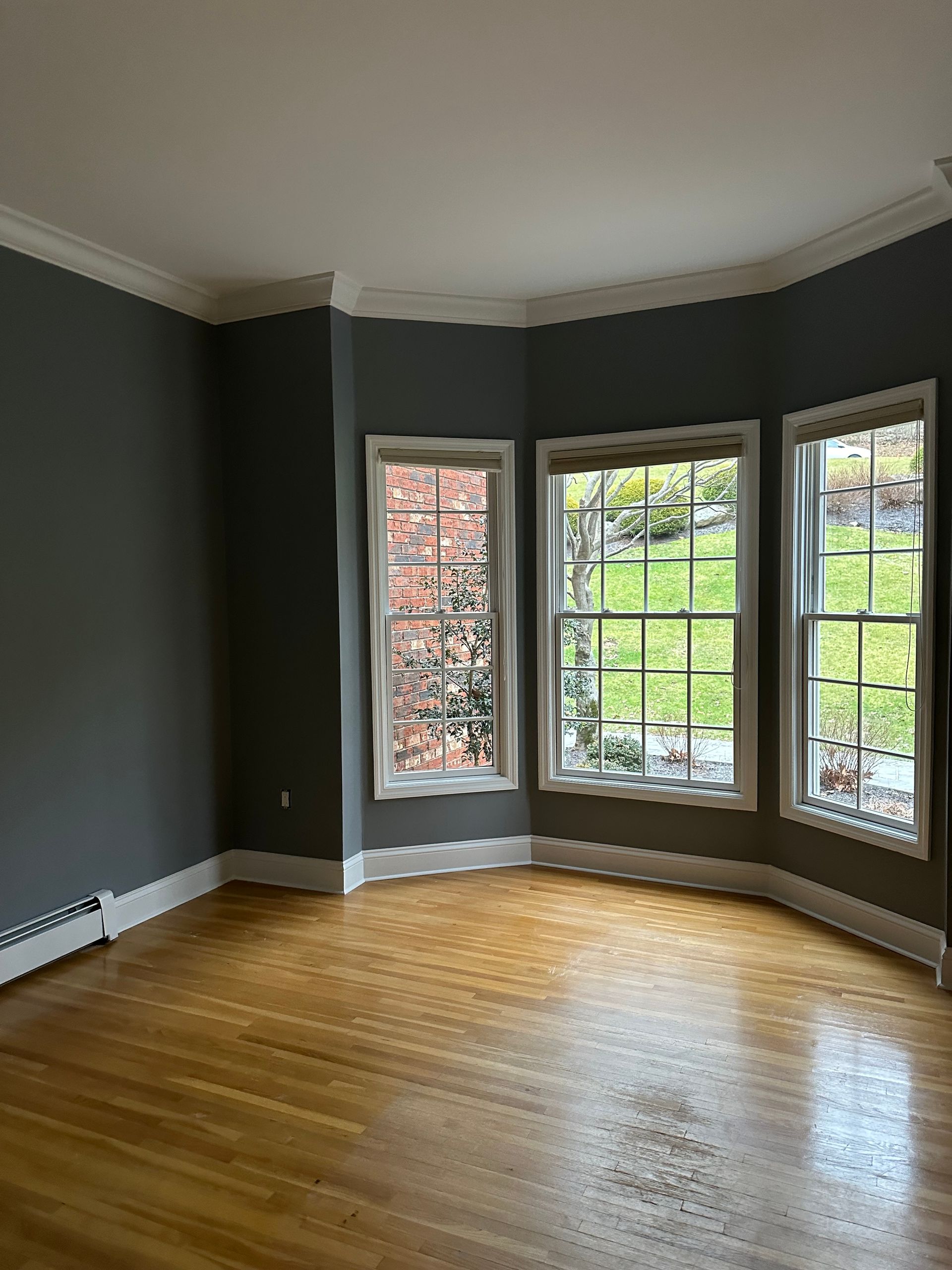 Empty room with hardwood floors, gray walls, and a curved bay window overlooking a green yard.