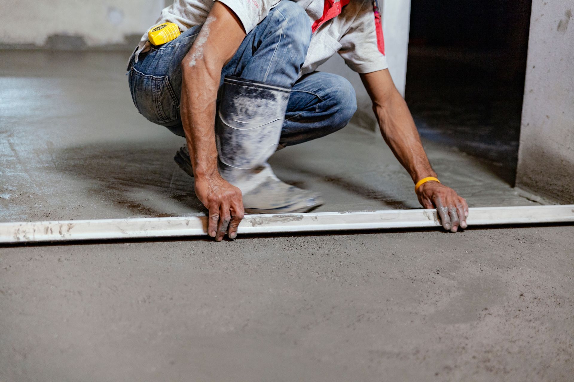 Person crouches, leveling wet concrete floor with a long straight edge, inside a room.