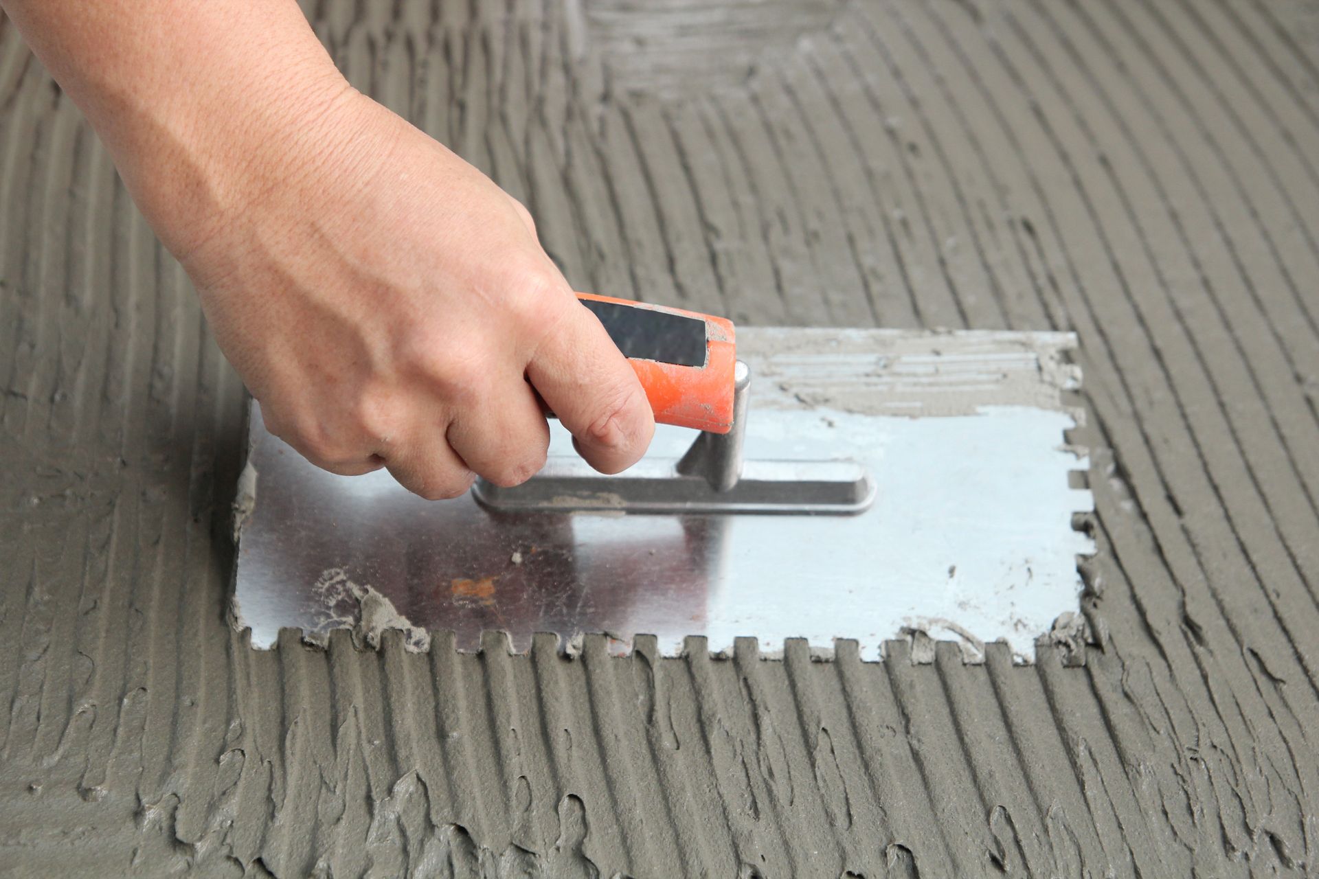 Hand using a notched trowel to spread gray mortar on a surface, creating ridges.