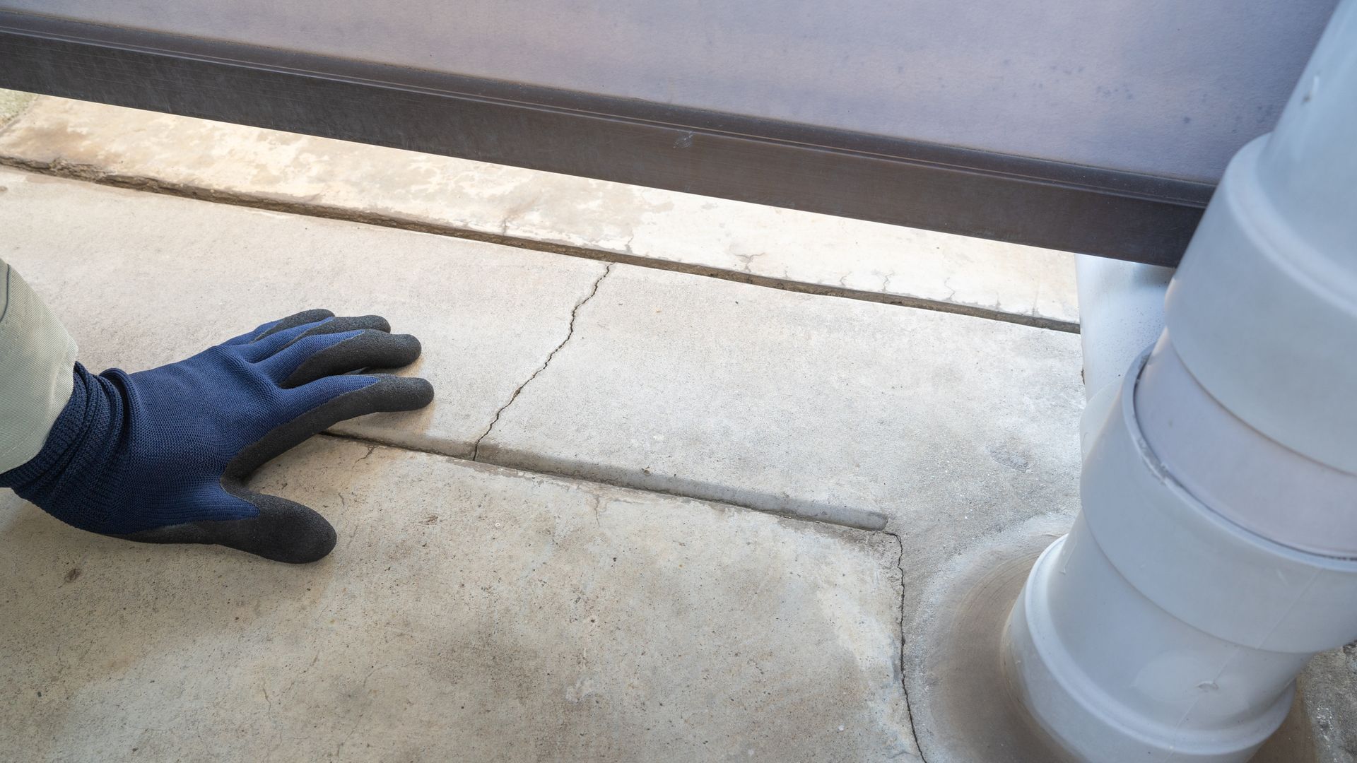 Gloved hand touching a concrete surface with a crack near a white pipe.