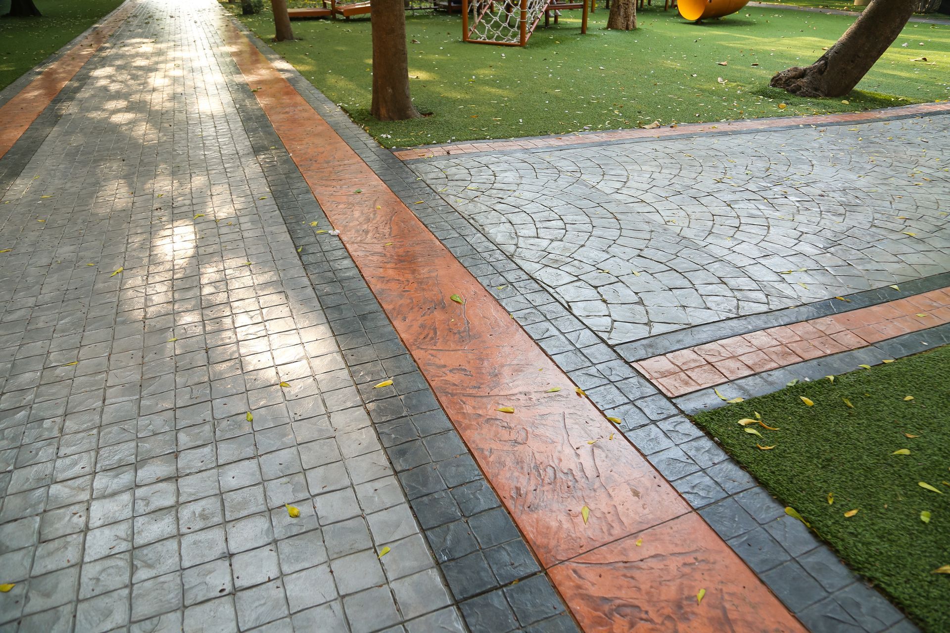 Paved walkway in a park with patterned gray and reddish-brown brickwork next to a green lawn.