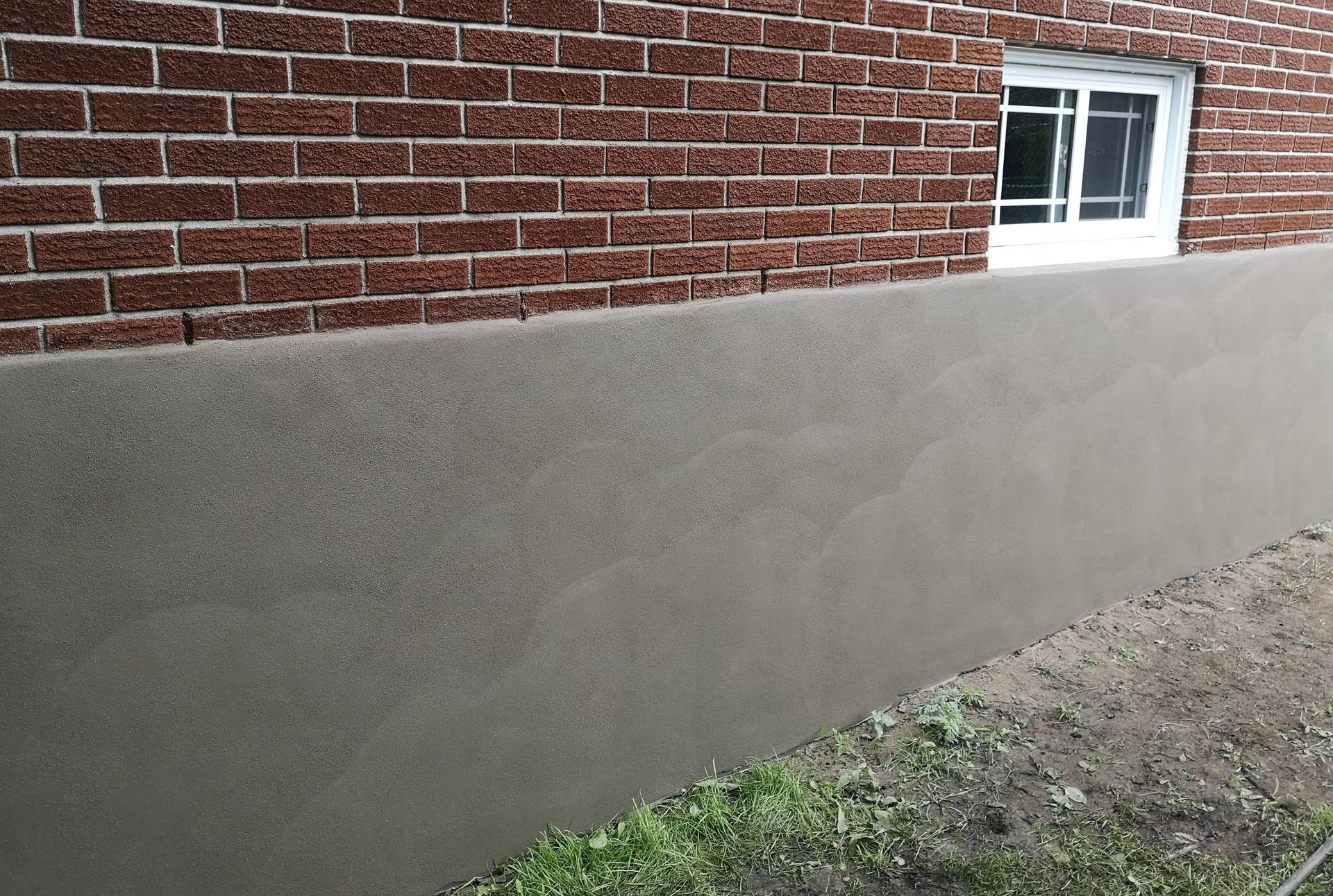 Brick wall with gray stucco foundation, small window, and grass in front.