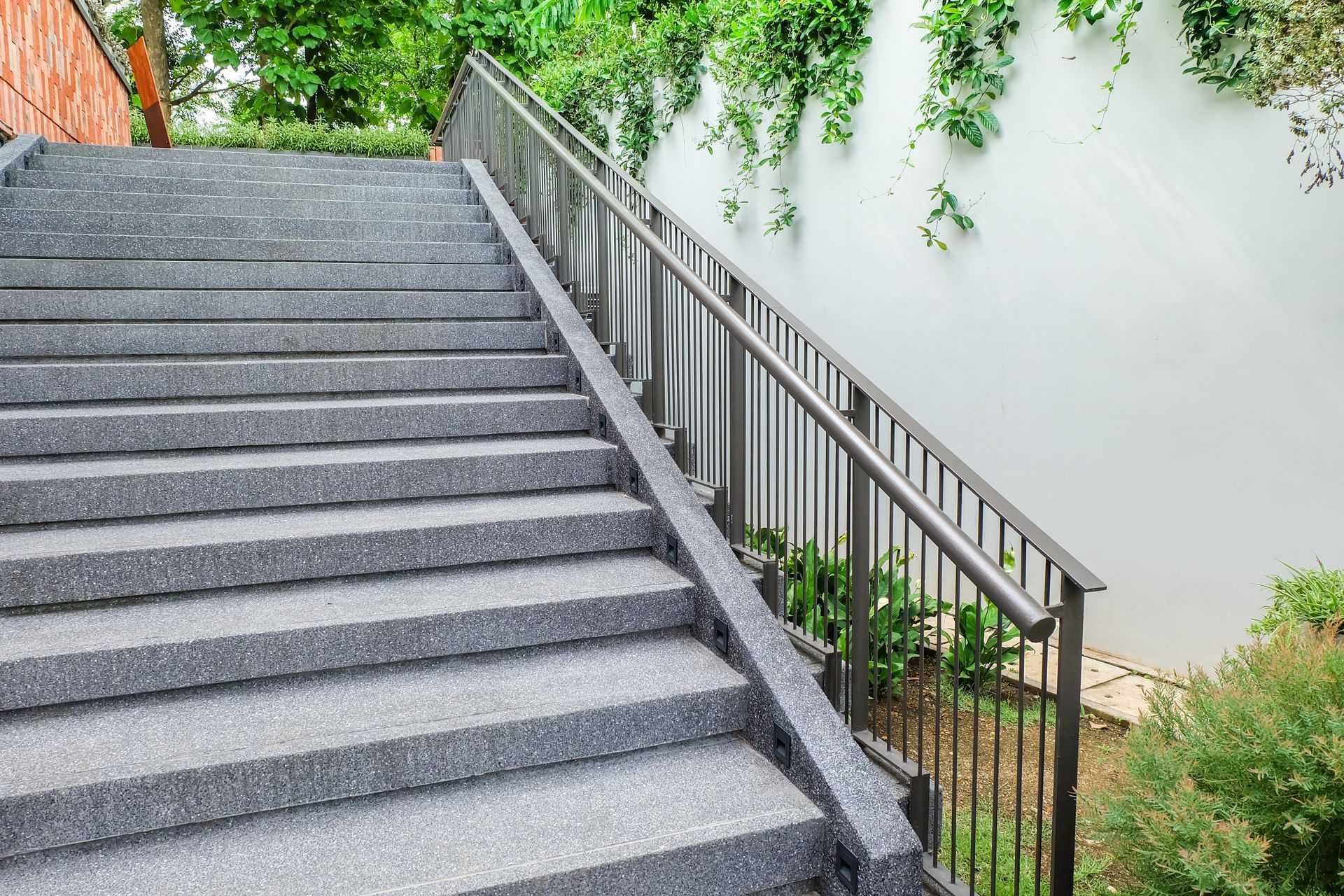 Gray stone steps with a metal handrail alongside a white wall with greenery.