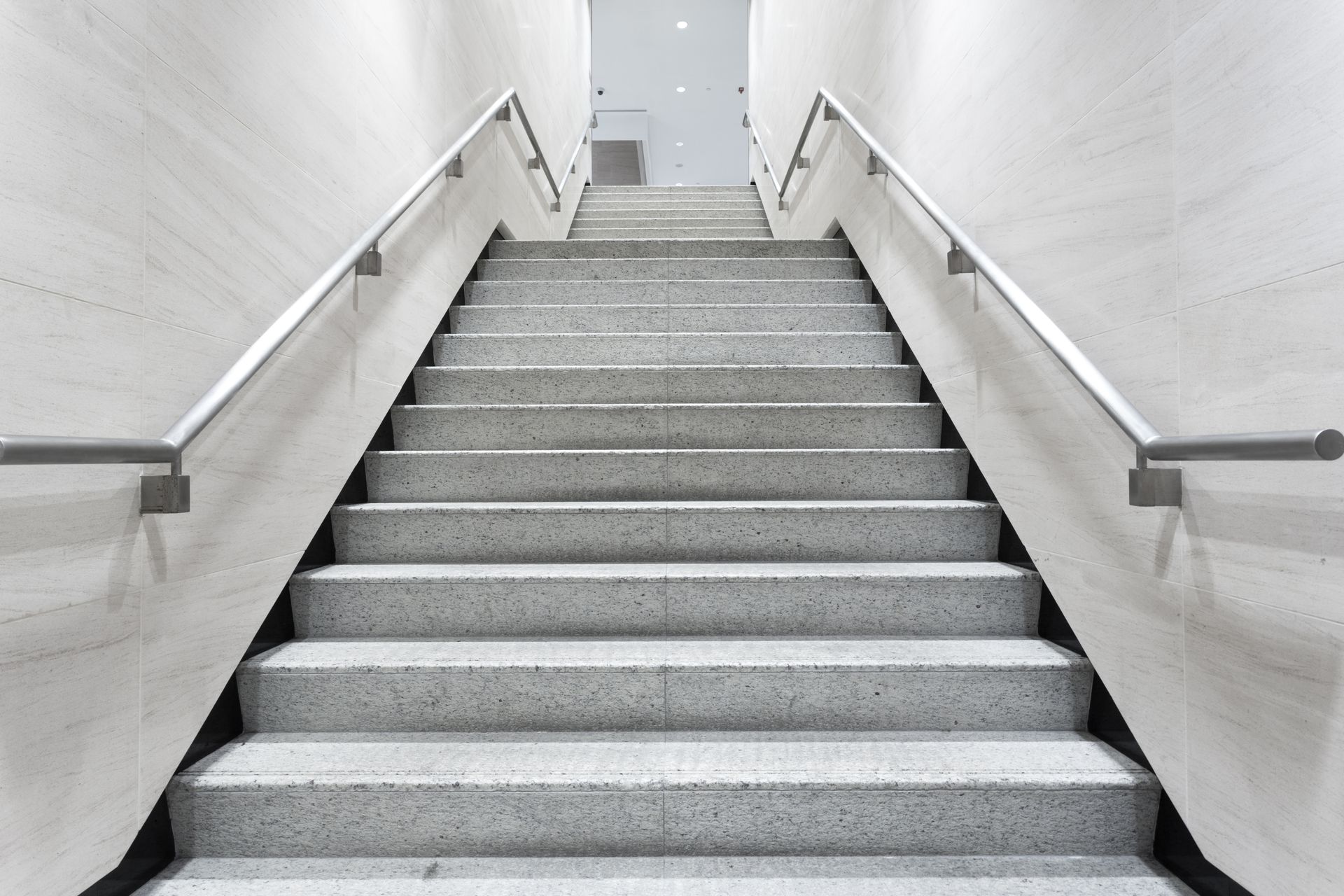 Staircase with gray stone steps and white walls, metal handrails on either side, leading upwards.