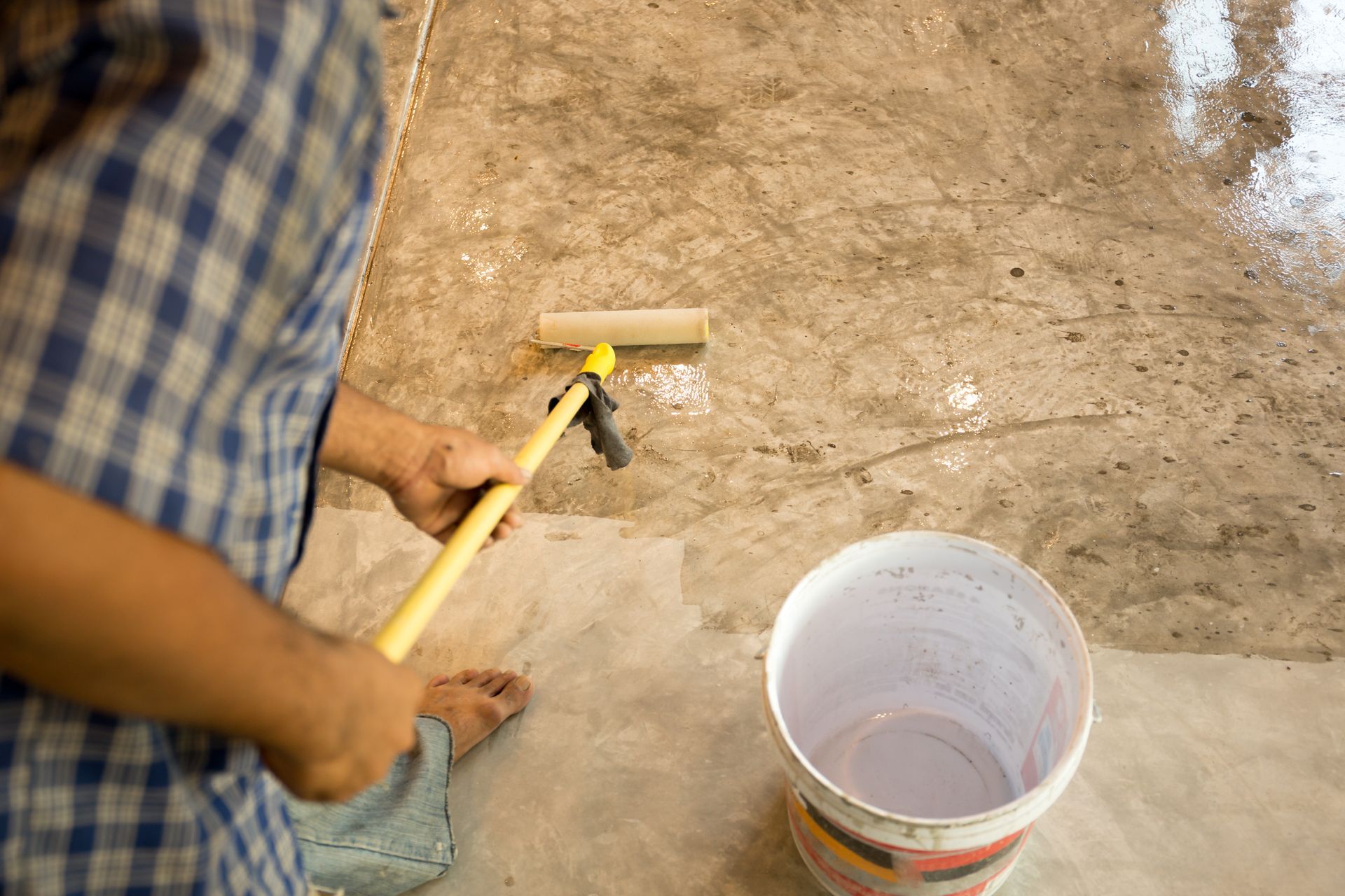 Person using a paint roller to coat a concrete floor with a white substance.