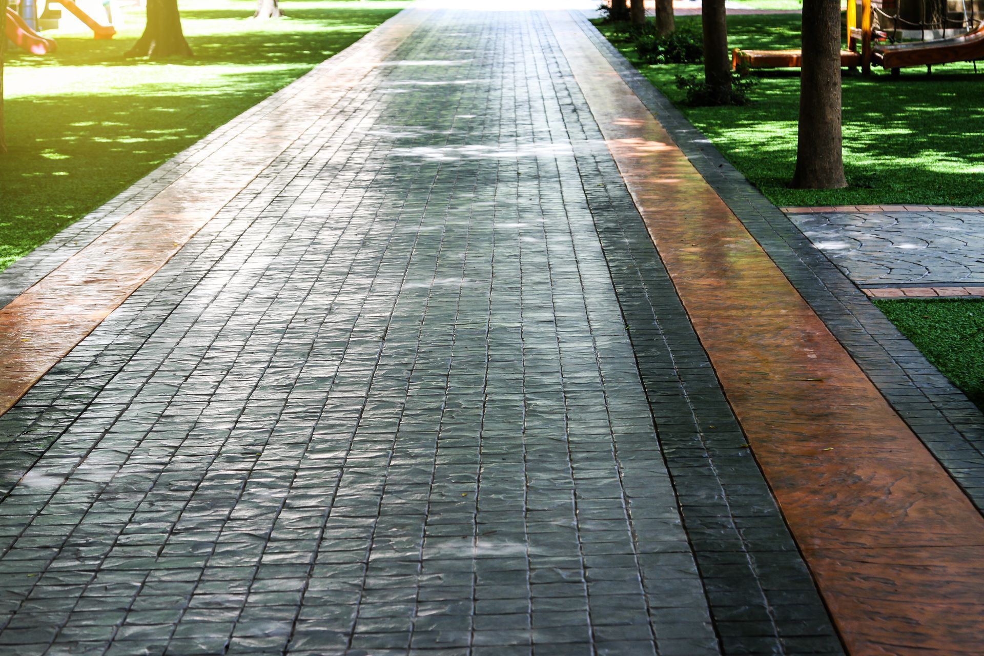Brick walkway with orange border through a park, dappled sunlight on the pavement.