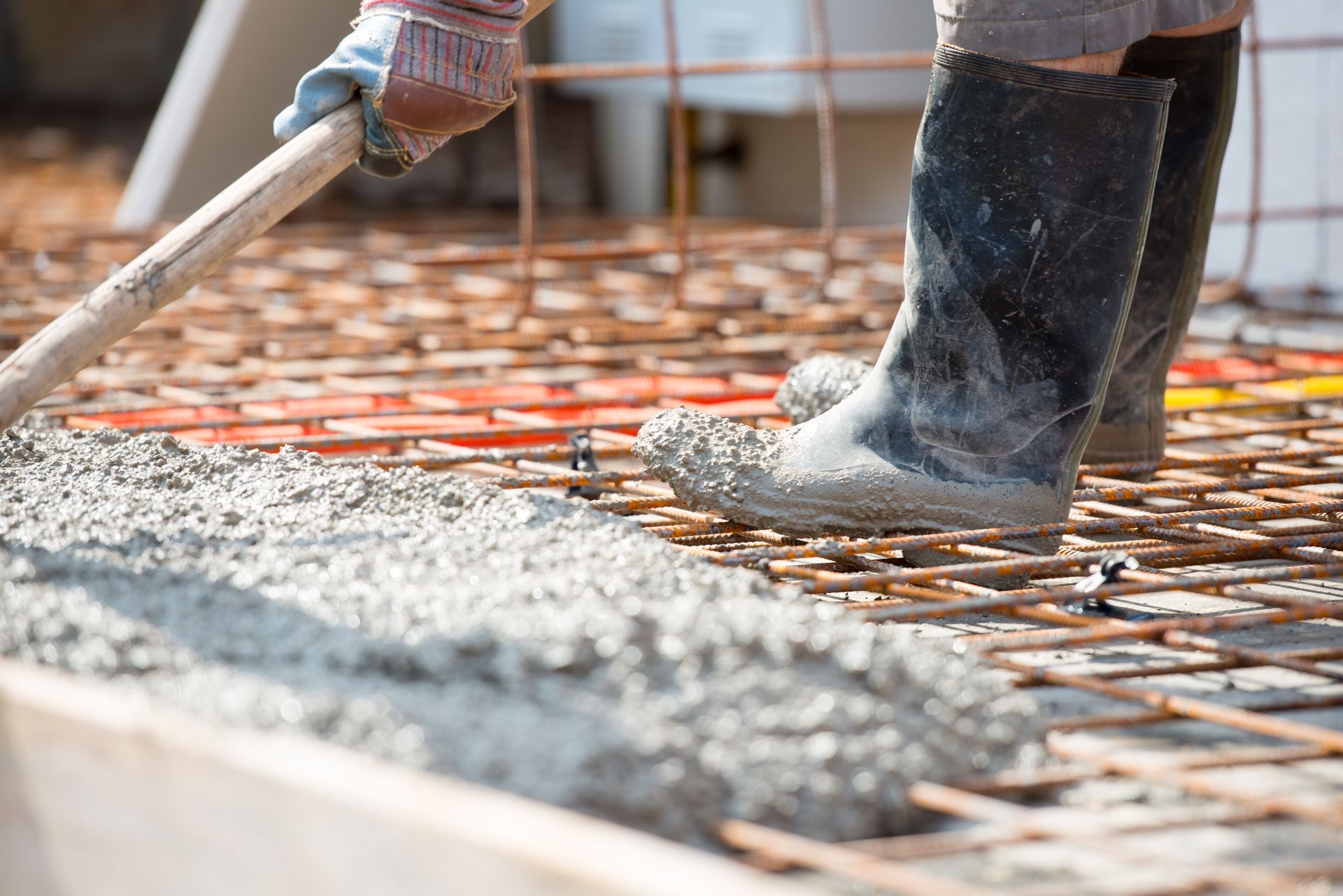 Worker in rubber boots spreading concrete over rebar with a wooden tool on a construction site.