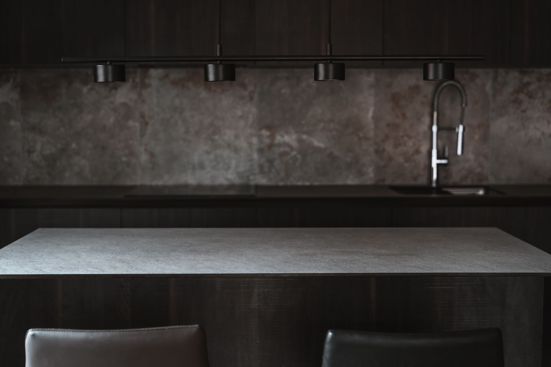 Dark modern kitchen with stone backsplash, island, sink, and pendant lights.