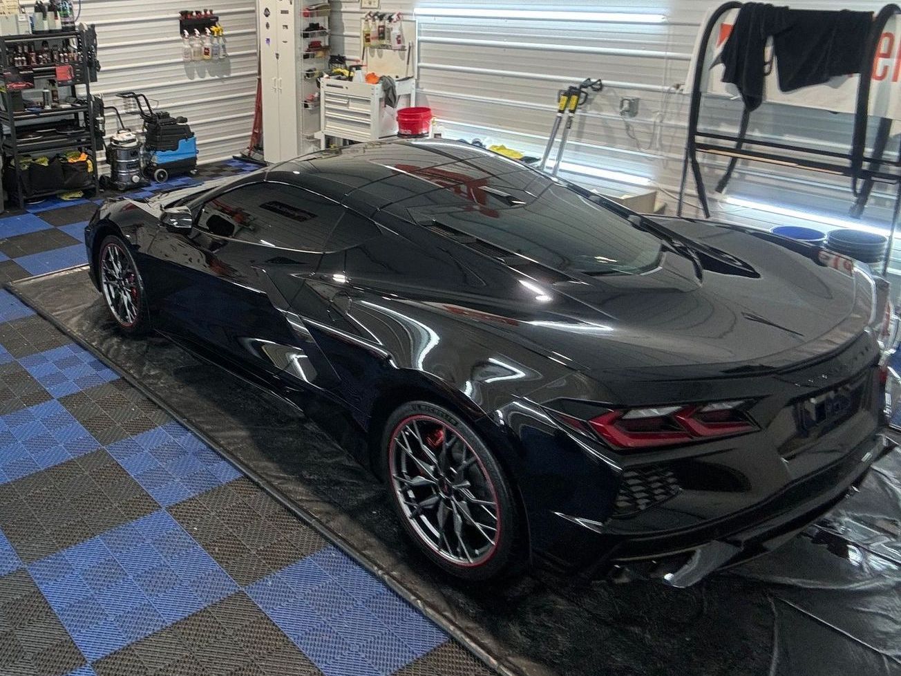 Black Corvette in a garage, shiny paint, red-trimmed wheels, blue and black floor tiles, detailing equipment.