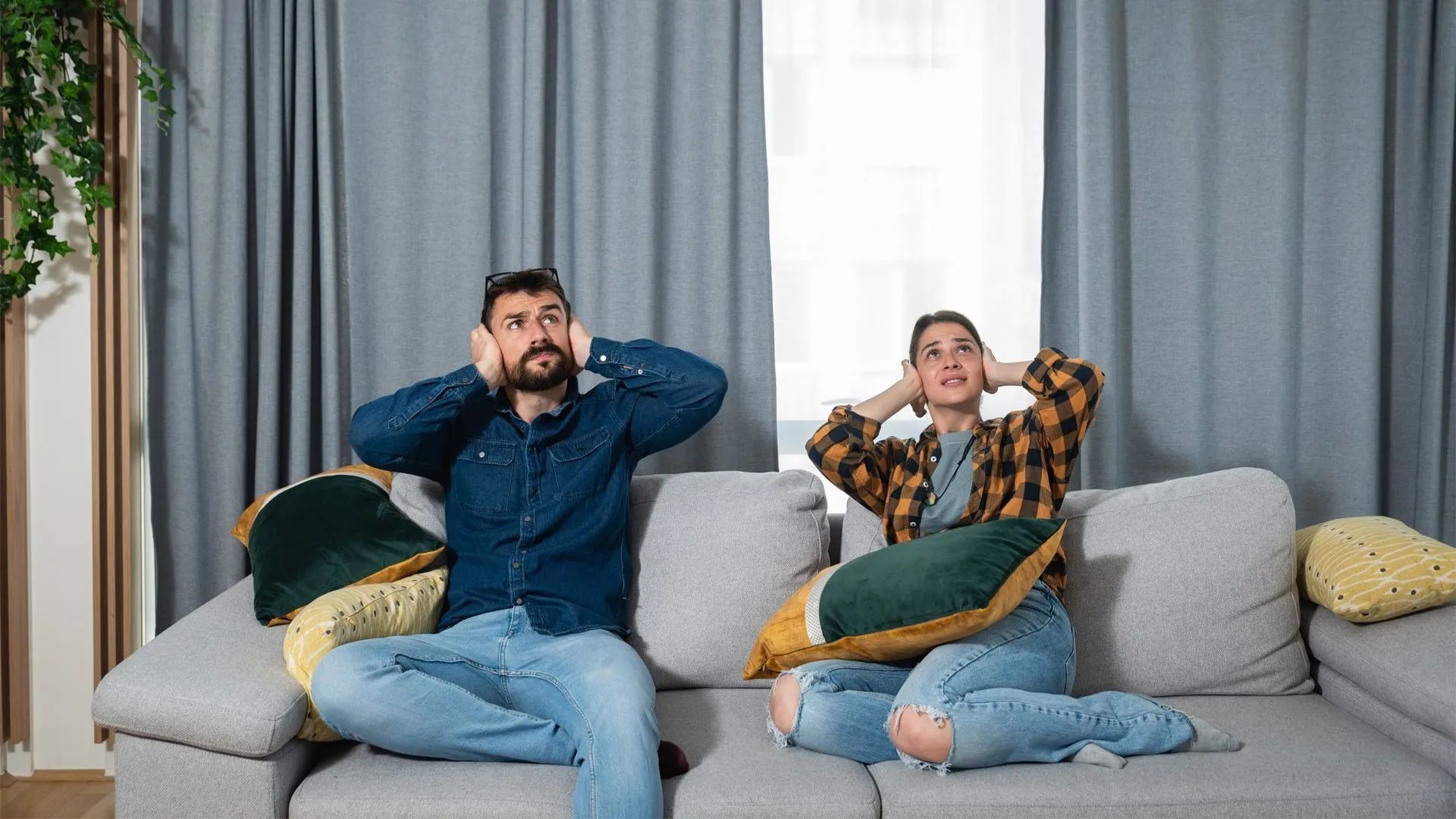 Couple sat on a sofa covering their ears to block out noise