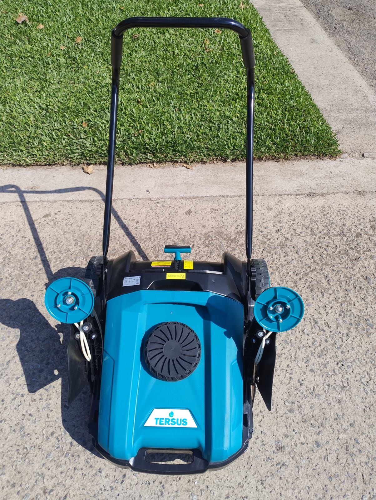 Un aireador de césped verde azulado y negro sobre una superficie pavimentada con césped al fondo.
