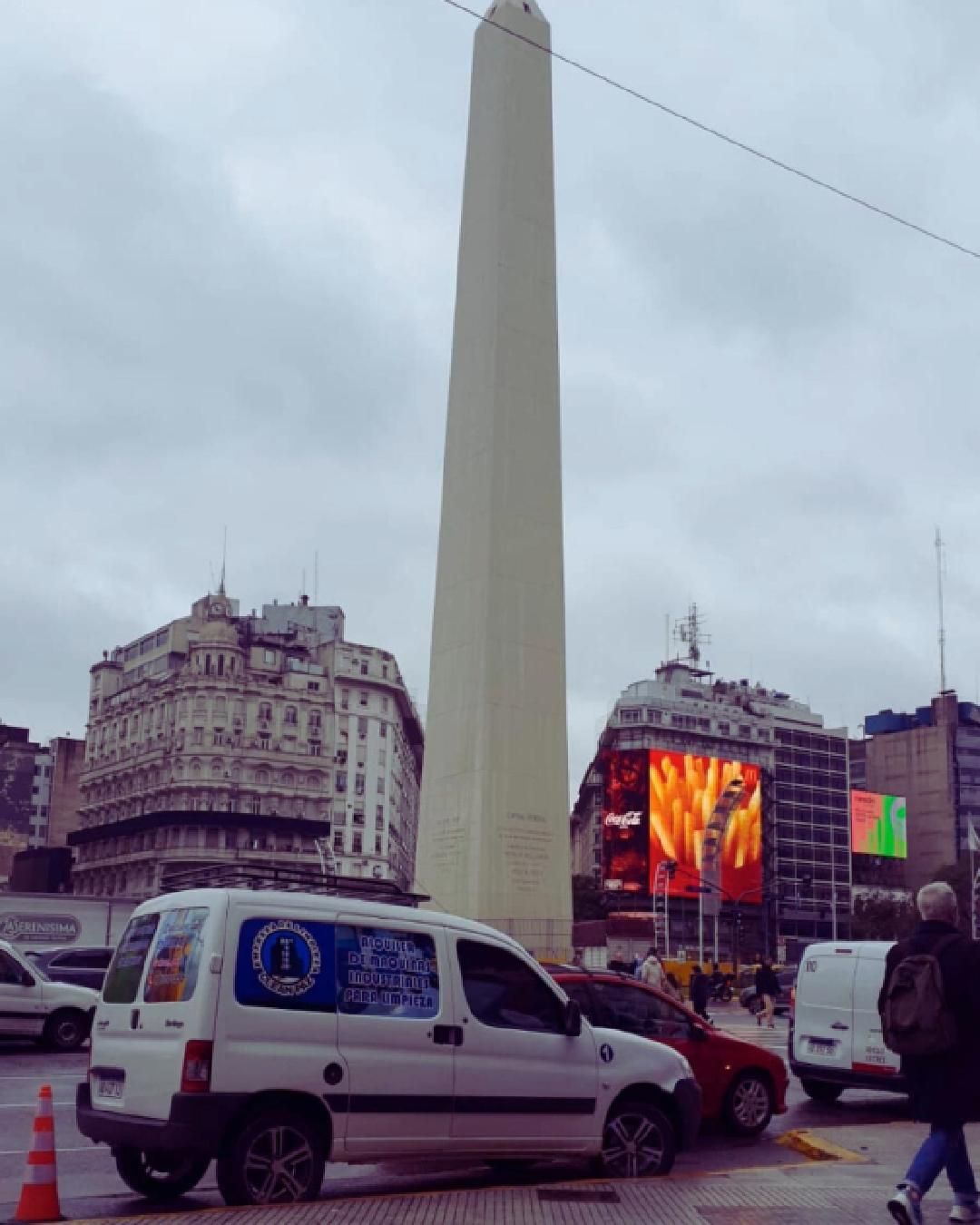 The Obelisk of Buenos Aires stands tall in a city square, surrounded by traffic, urban buildings, and digital billboards.