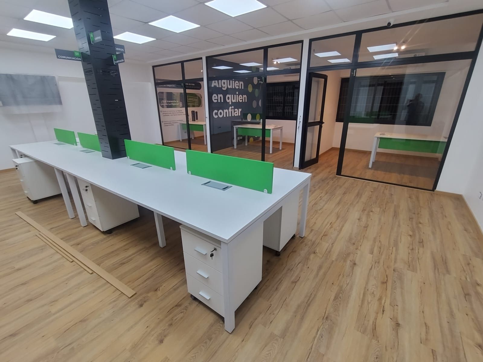 Interior de oficina con largos escritorios blancos, divisores verdes, paredes de vidrio y pisos de madera.