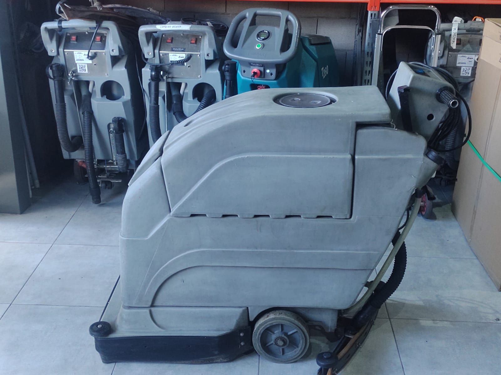 Máquina de limpieza de suelos gris, con varias máquinas al fondo. Entorno industrial.