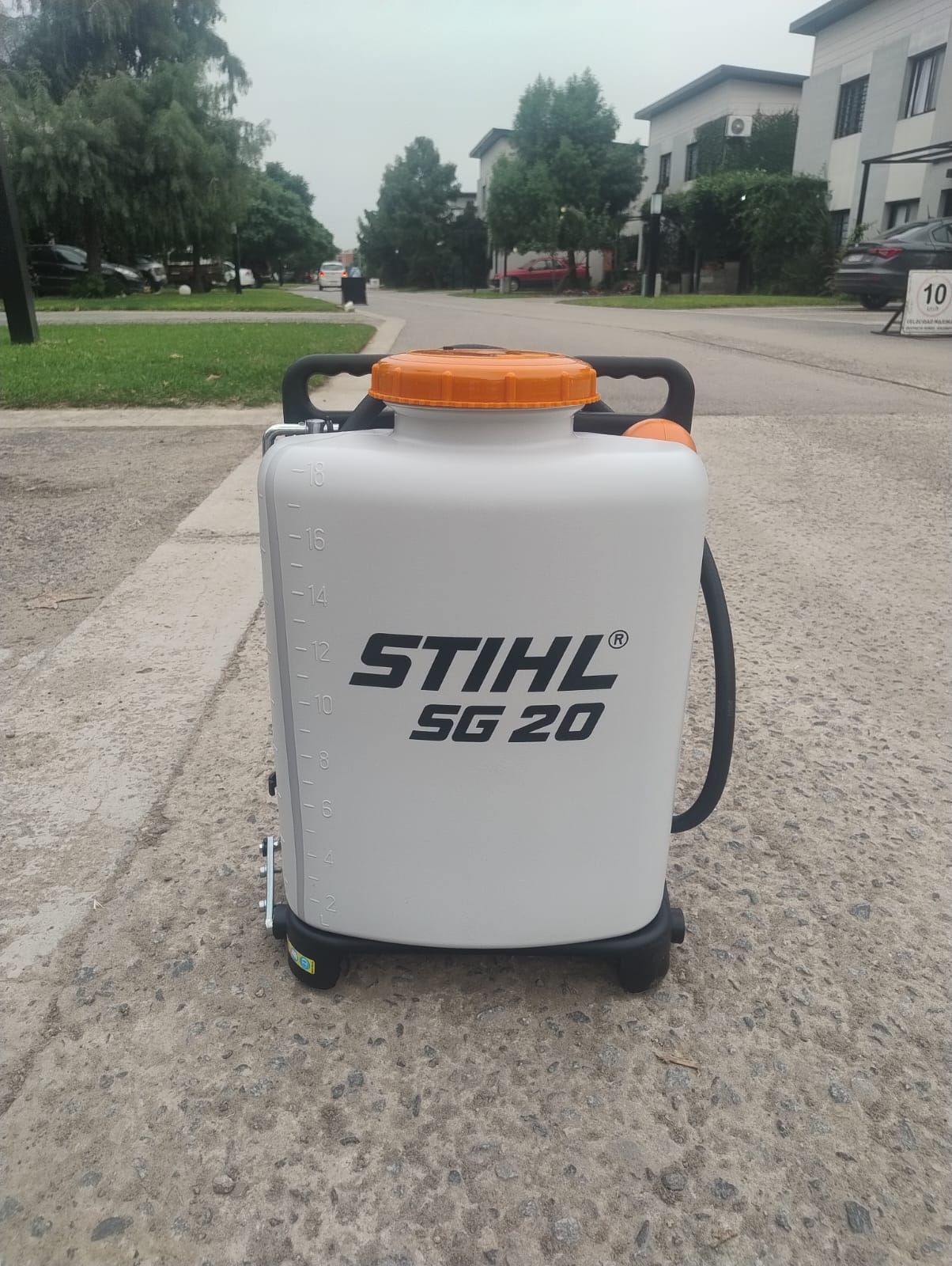 Pulverizador de mochila STIHL SG 20 de color blanco con parte superior naranja, sobre una acera gris, en una zona suburbana.