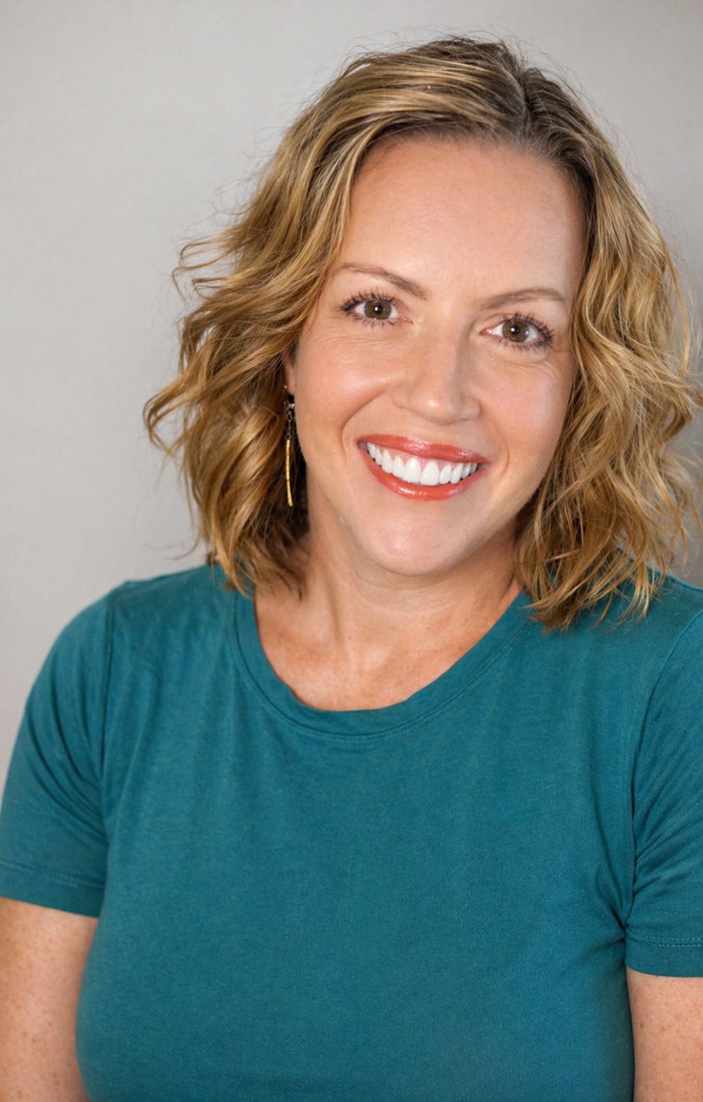 A woman in a tan coat is smiling for the camera in a park.