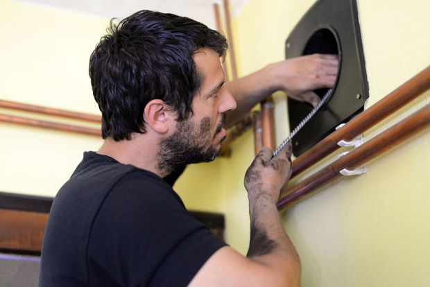 Un uomo, con il viso e le mani ricoperti di fuliggine, pulisce il camino, il muro giallo e i tubi di rame.