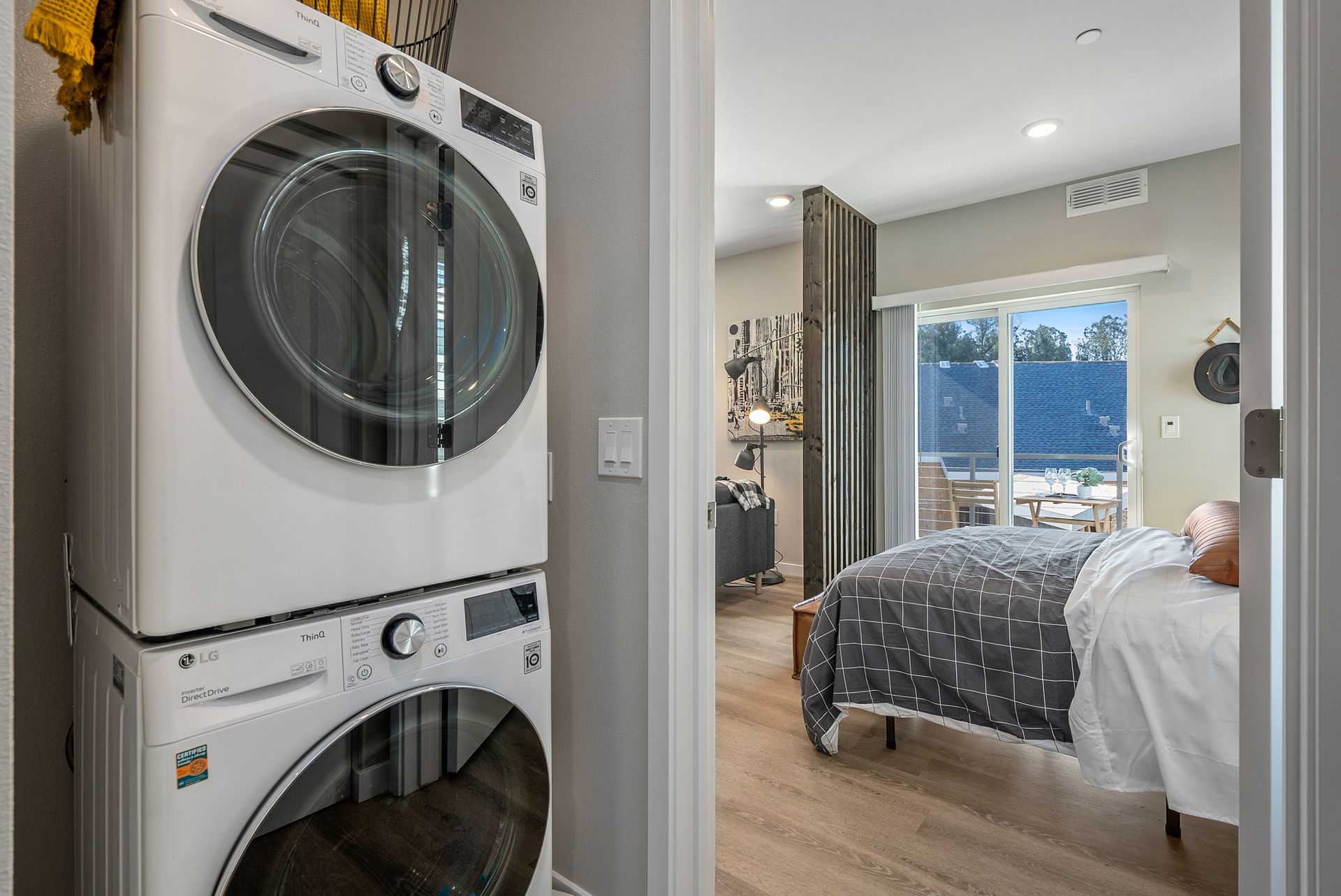 Stacked white washer and dryer in a laundry room, doorway leads to a bedroom with a bed and balcony view.