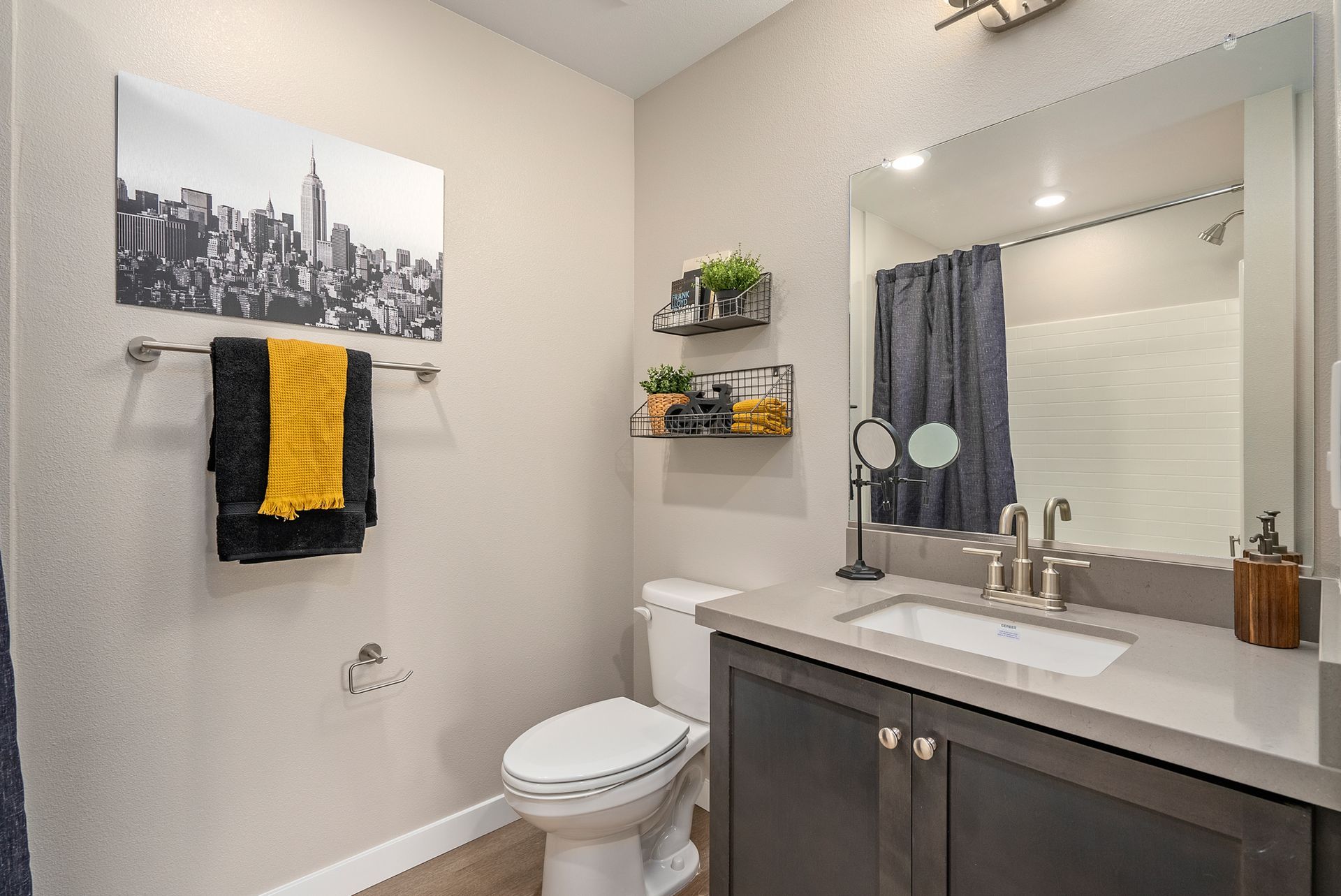 Bathroom with a grey vanity, white toilet, and black and yellow towels. NYC skyline art and green plants.