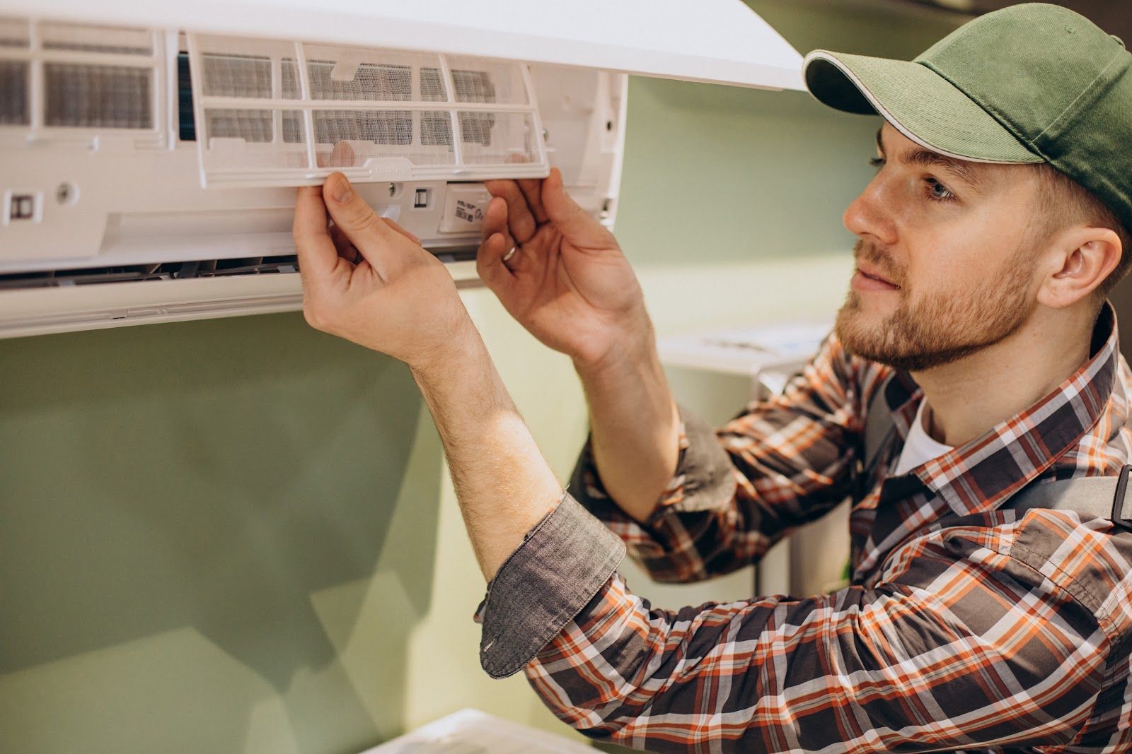Man in plaid shirt and cap cleaning an air conditioner filter, indoors.