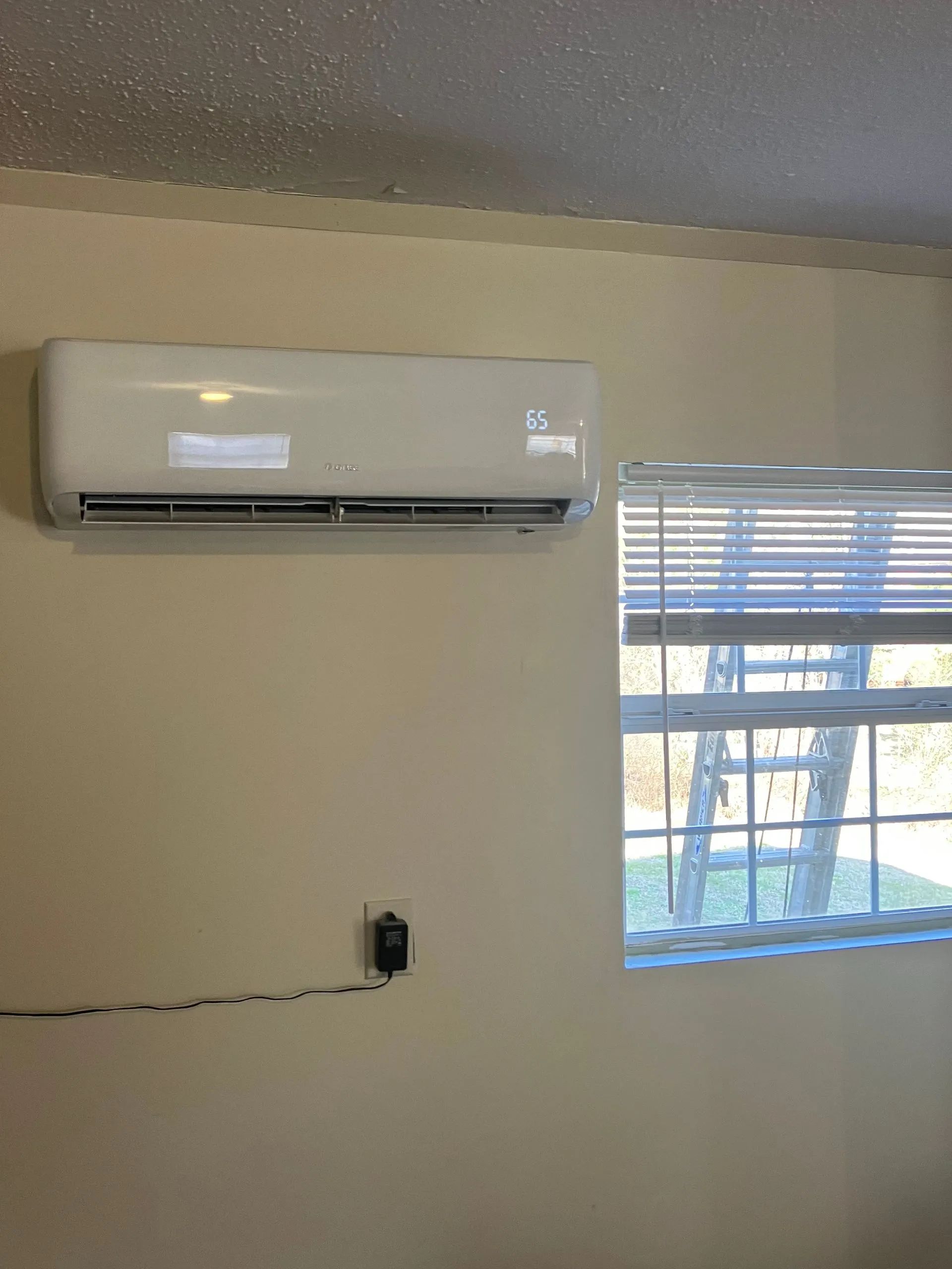 Wall-mounted air conditioner above a window with blinds in a beige room, with a wire outlet on the wall.