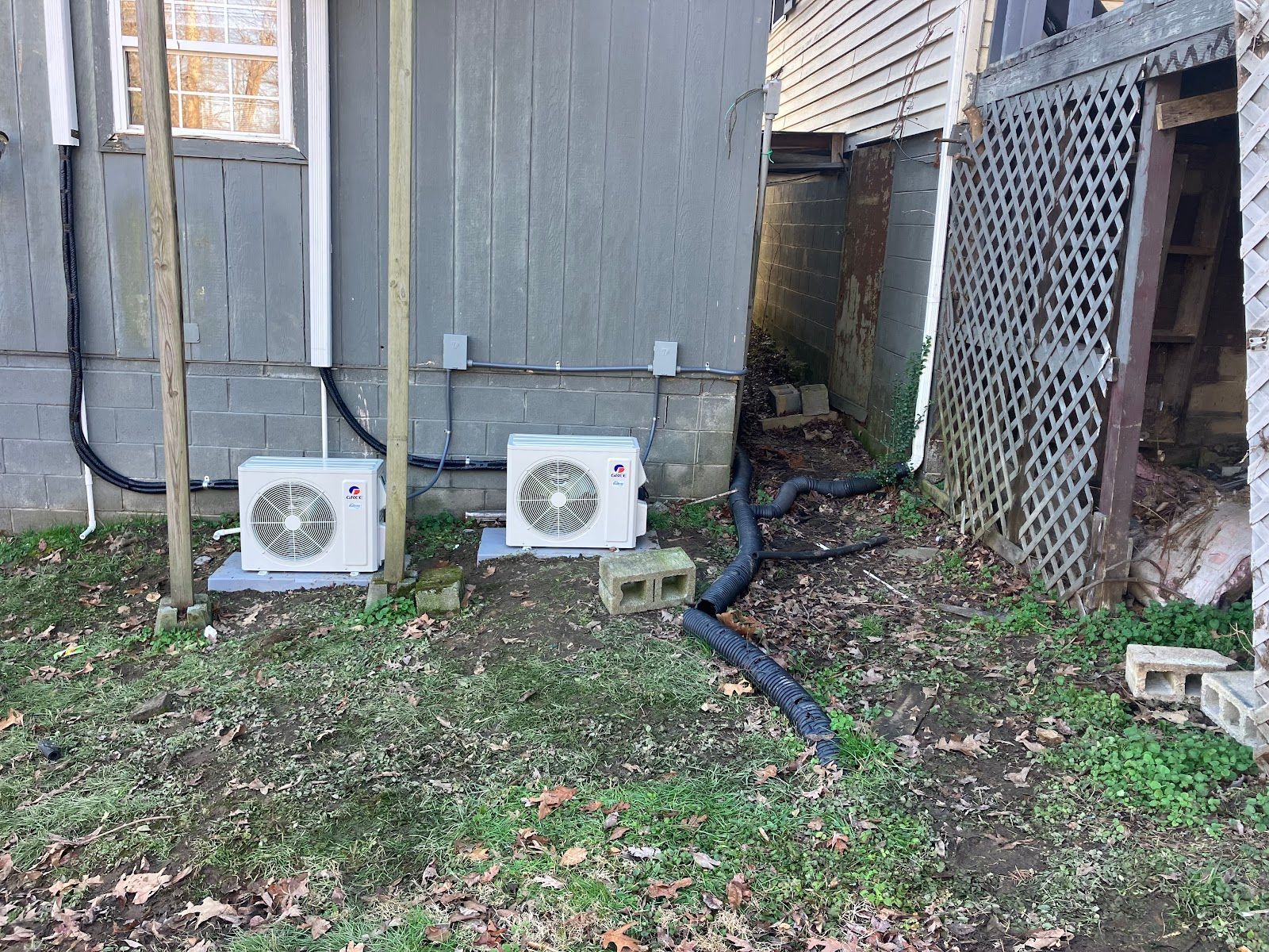 Two white air conditioning units outside a gray building, with black tubing and drainage.