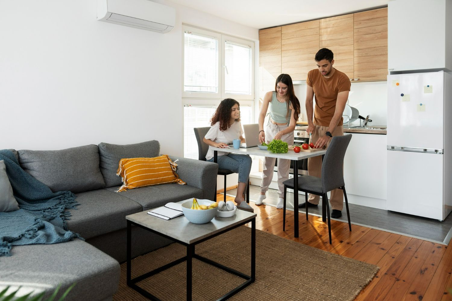 Family in a small kitchen/living area. Mother and father prepare food while daughter works at table.