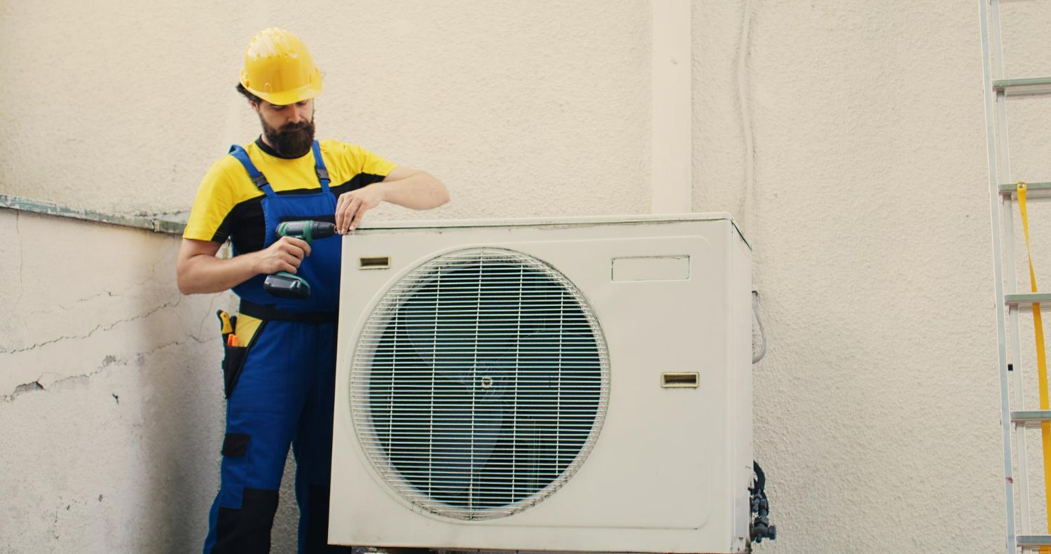A repairman in overalls and a hard hat uses a drill on an air conditioning unit.