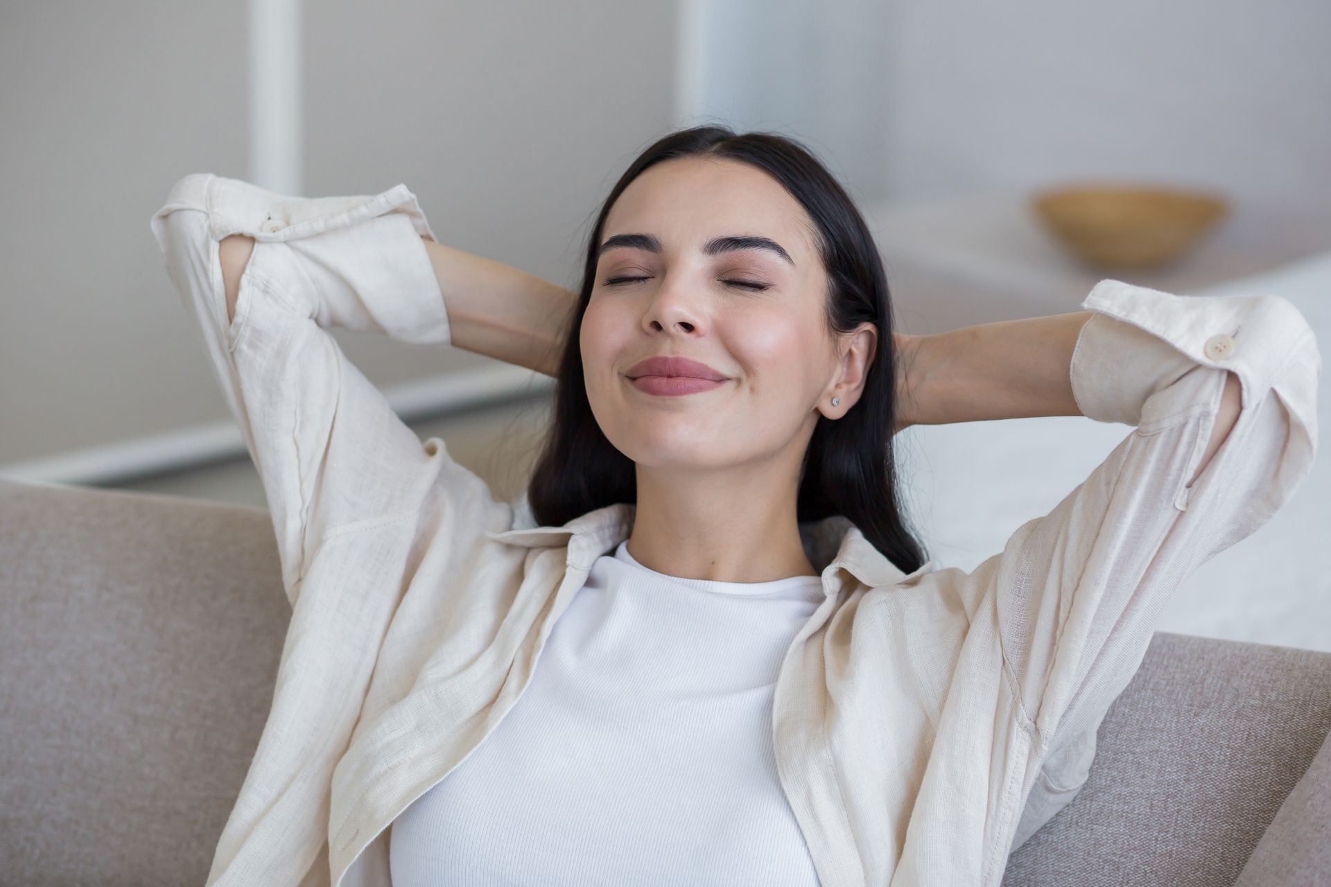 A woman is sitting on a couch with her eyes closed and her hands behind her head.