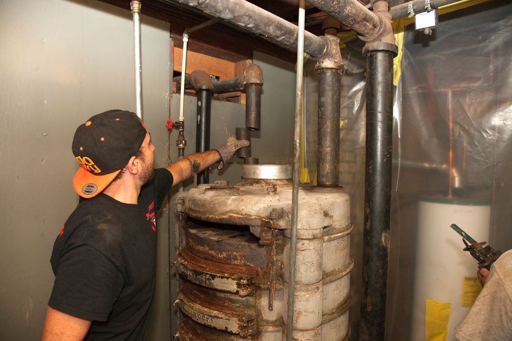 Man in black shirt points to a large, rusted industrial water heater in a utility room.
