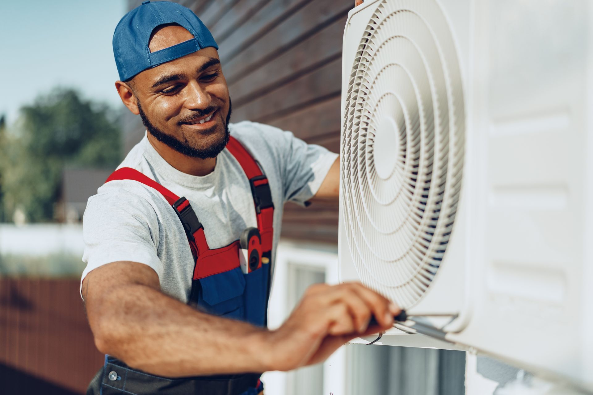 Technician in blue cap and coveralls, smiling, working on an AC unit outside a house.