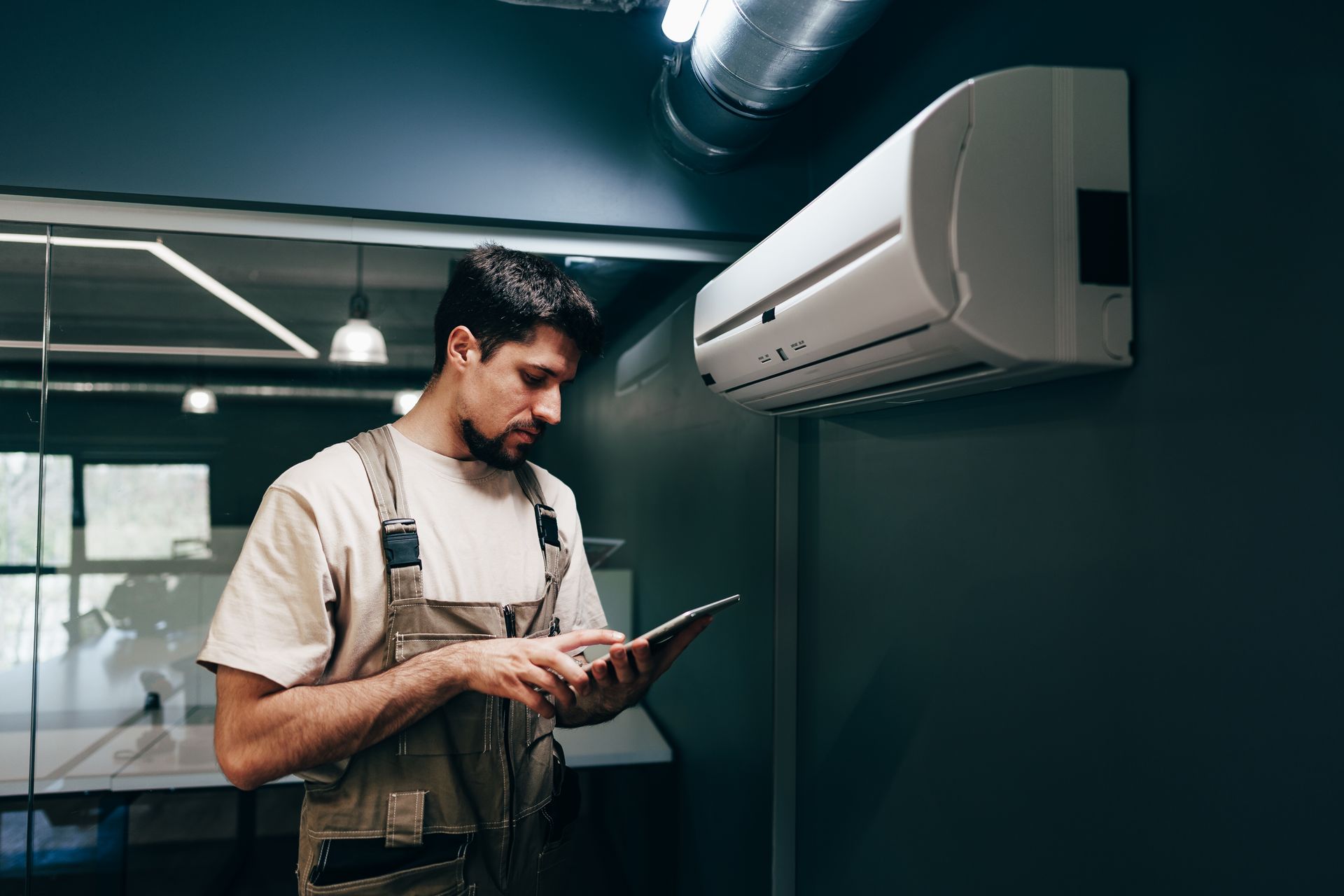 HVAC technician inspecting mini-split air conditioner unit with a tablet in a dark room.