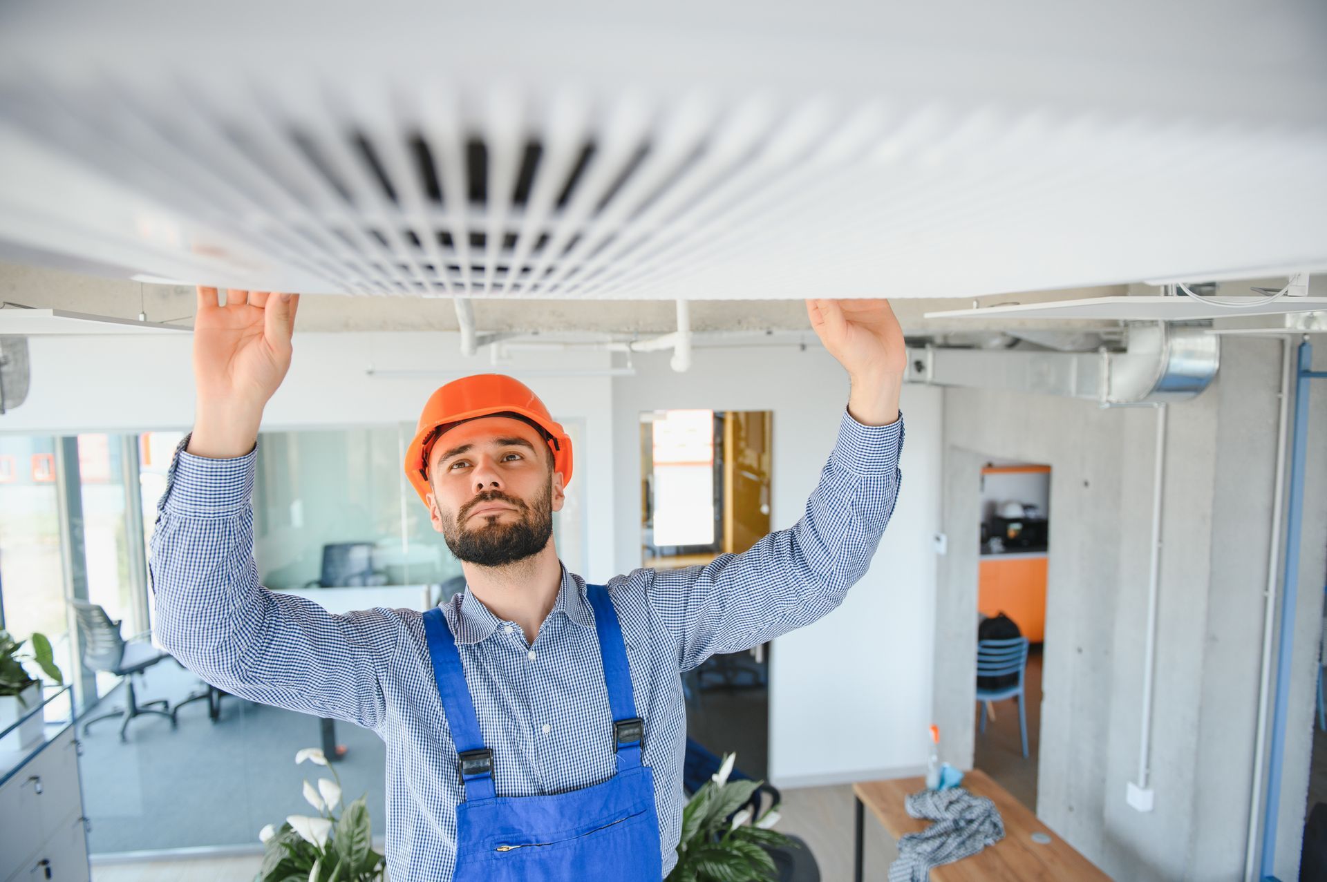 Man in blue overalls and hard hat inspecting ceiling ventilation in an office.