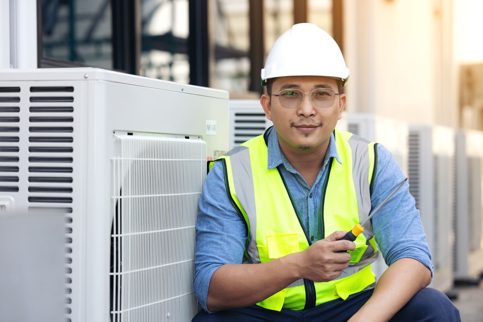 A man wearing a hard hat and safety vest is sitting next to an air conditioner.