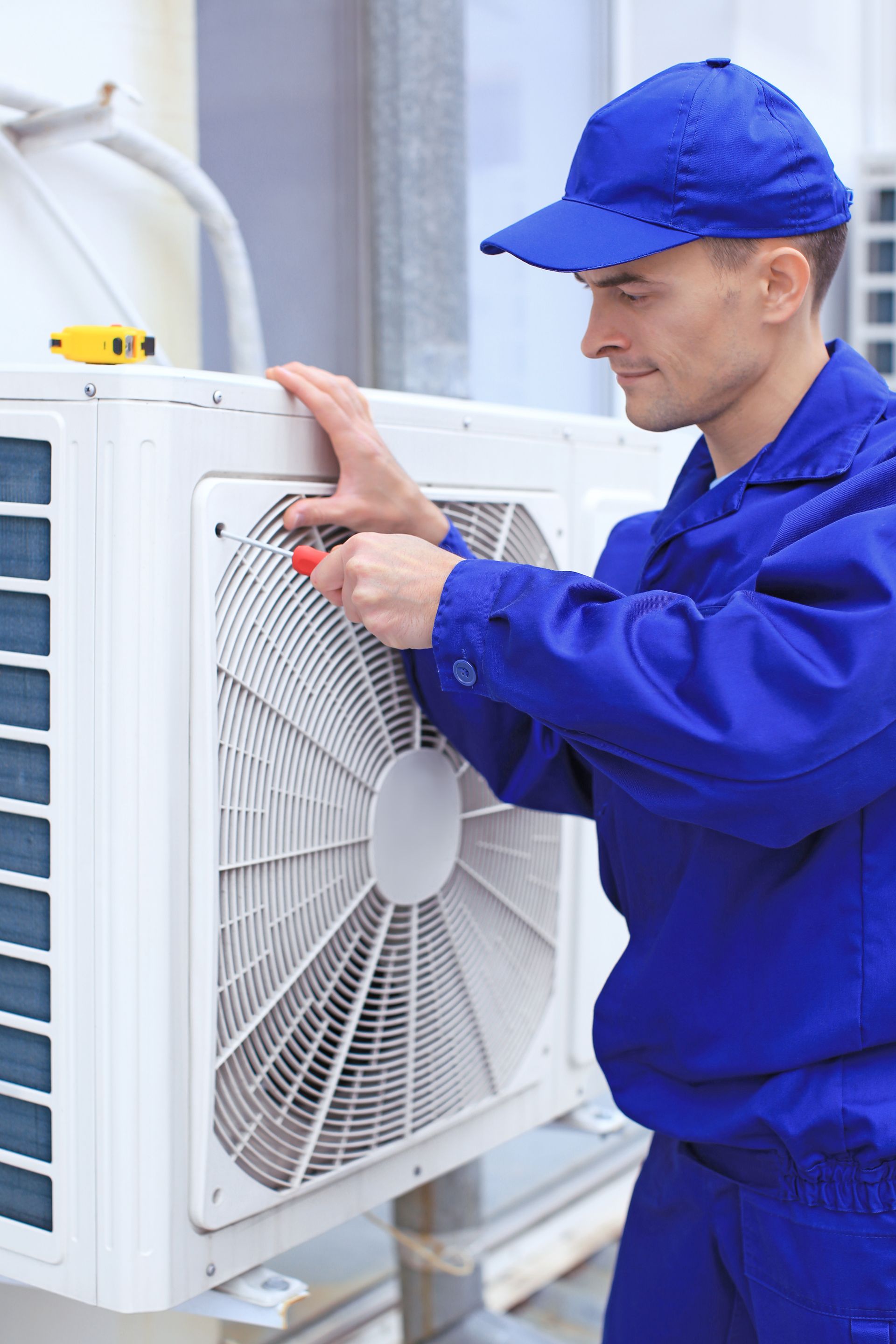 A man in a blue uniform is fixing an air conditioner with a screwdriver.