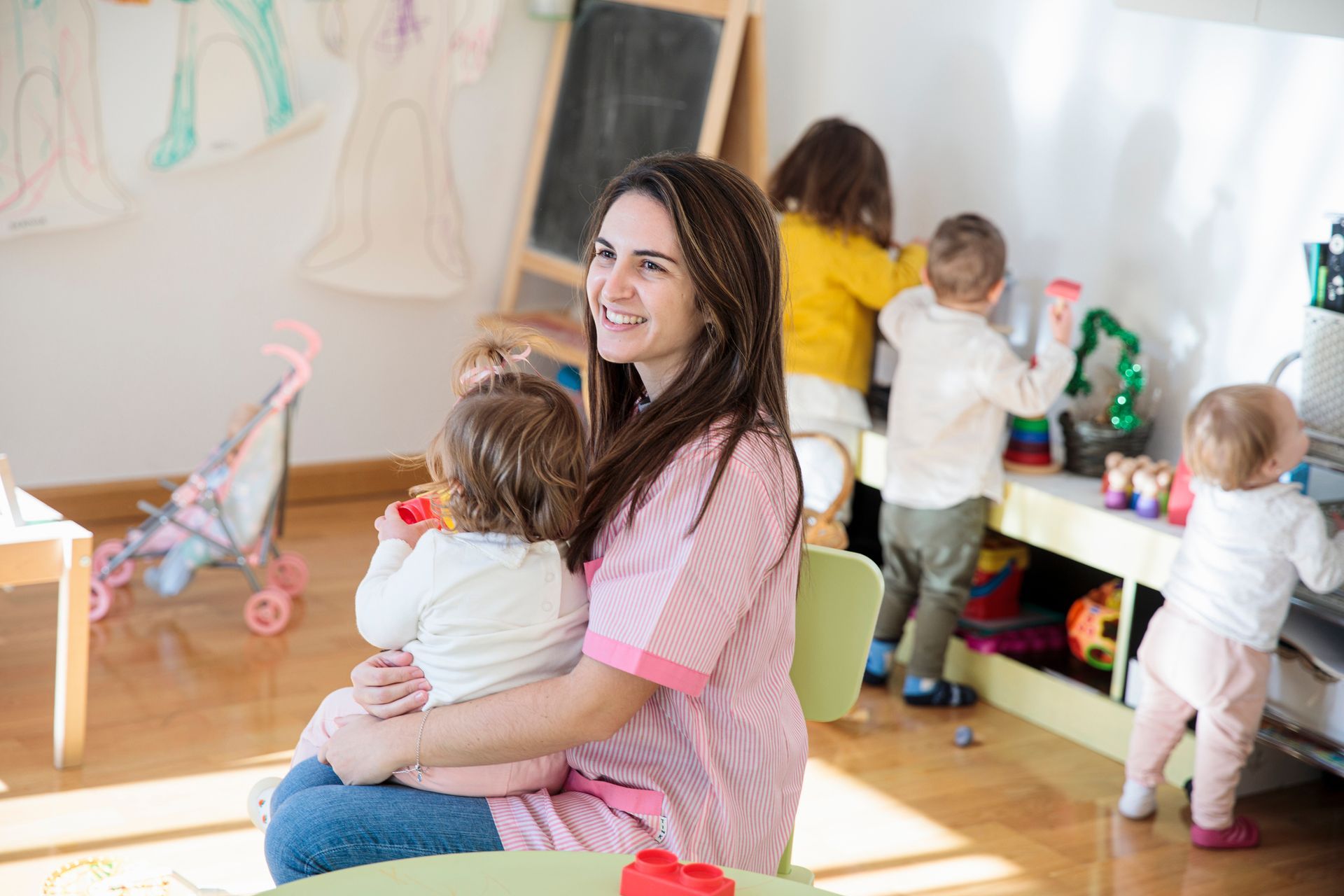 Female education teacher wearing pink t-shirt with a little girl in her arms at a toddler daycare.