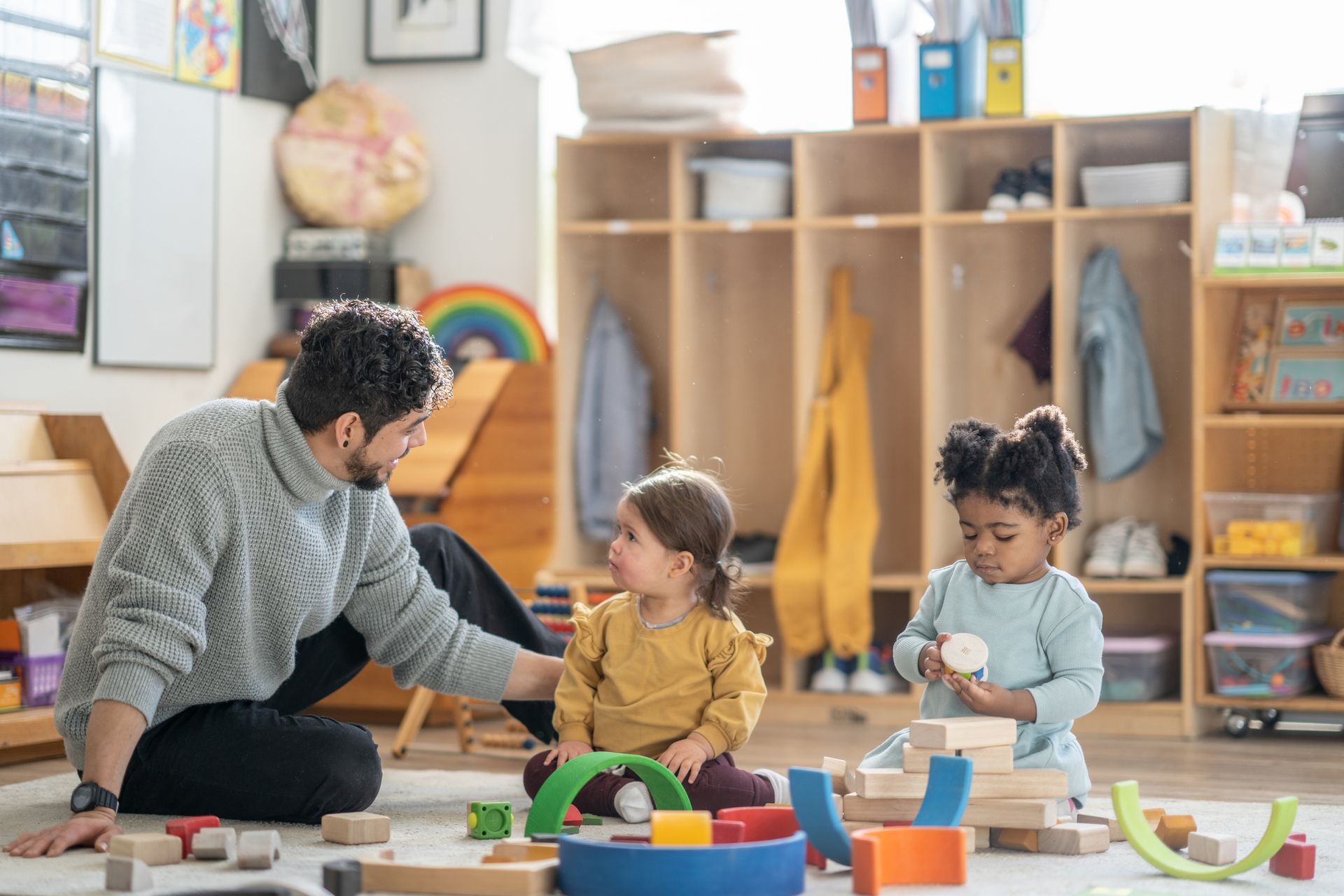 Daycare worker and children sit on the floor as the child plays with blocks.