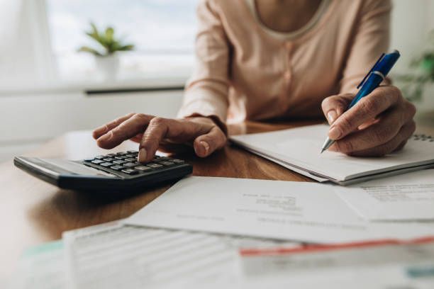 A woman is sitting at a table using a calculator and writing in a notebook.