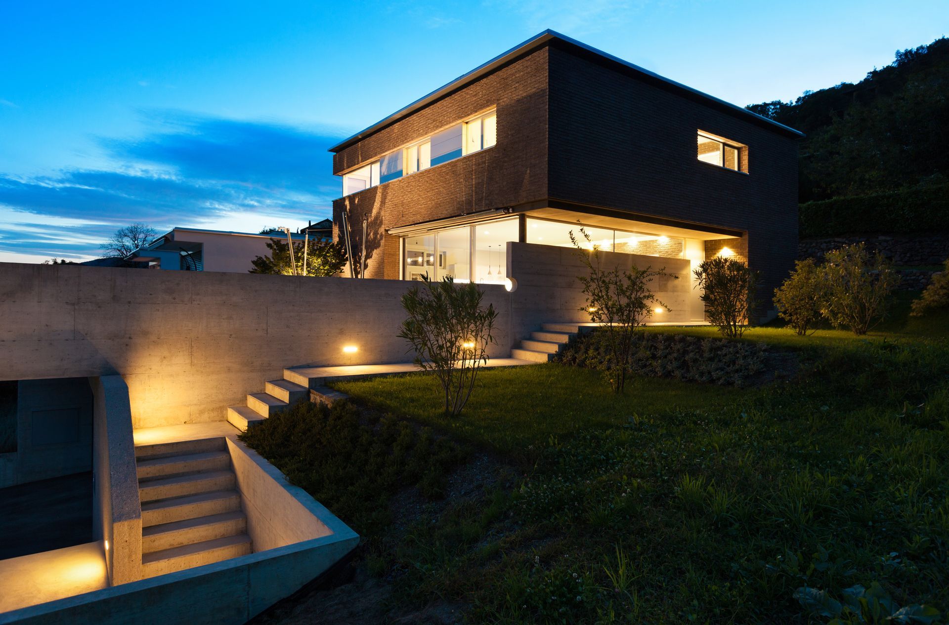 Modern two-story house at dusk, with illuminated interior and exterior lights, concrete features, and grassy yard.