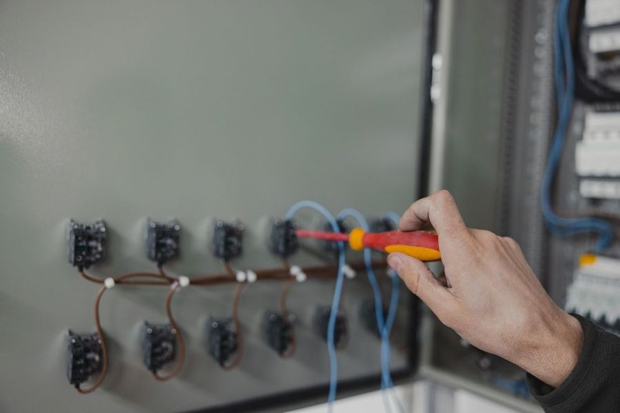 Person using a screwdriver to work on electrical wiring inside a gray control panel.
